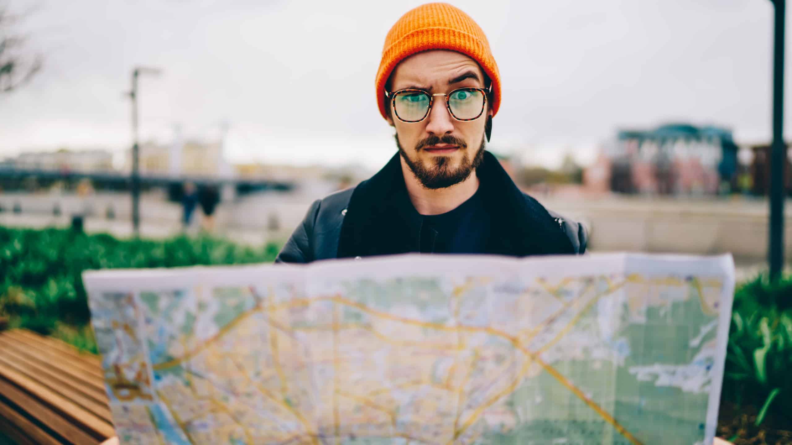 Portrait of puzzled tourist 20s holding paper location map confused with lost direction and destination, Caucasian hipster guy in trendy spectacles looking at camera during city sightseeing