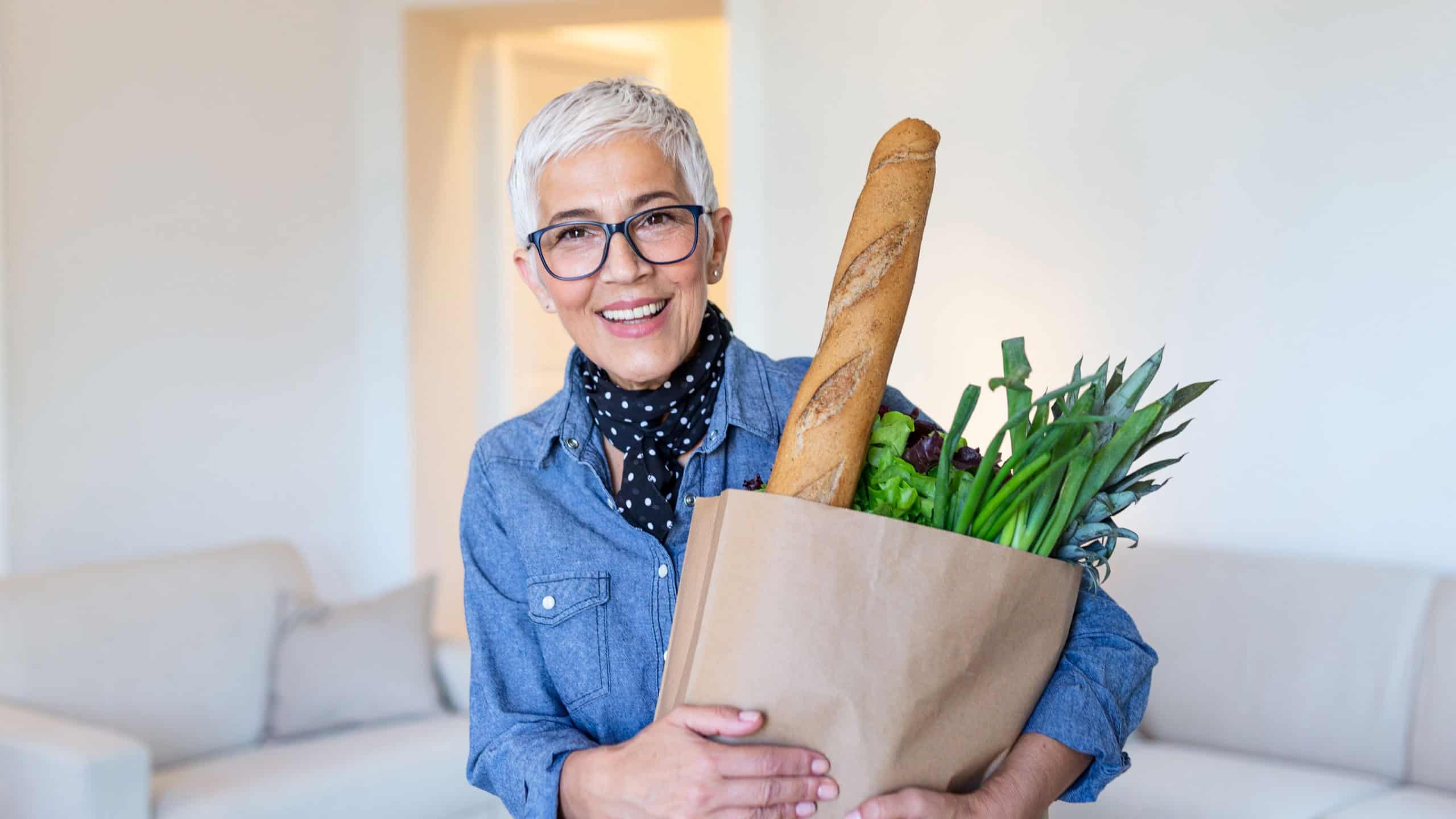 Healthy positive happy woman holding a paper shopping bag full of fruit and vegetables. Portrait of beautiful senior woman grocery shopping bag with vegetables at home
