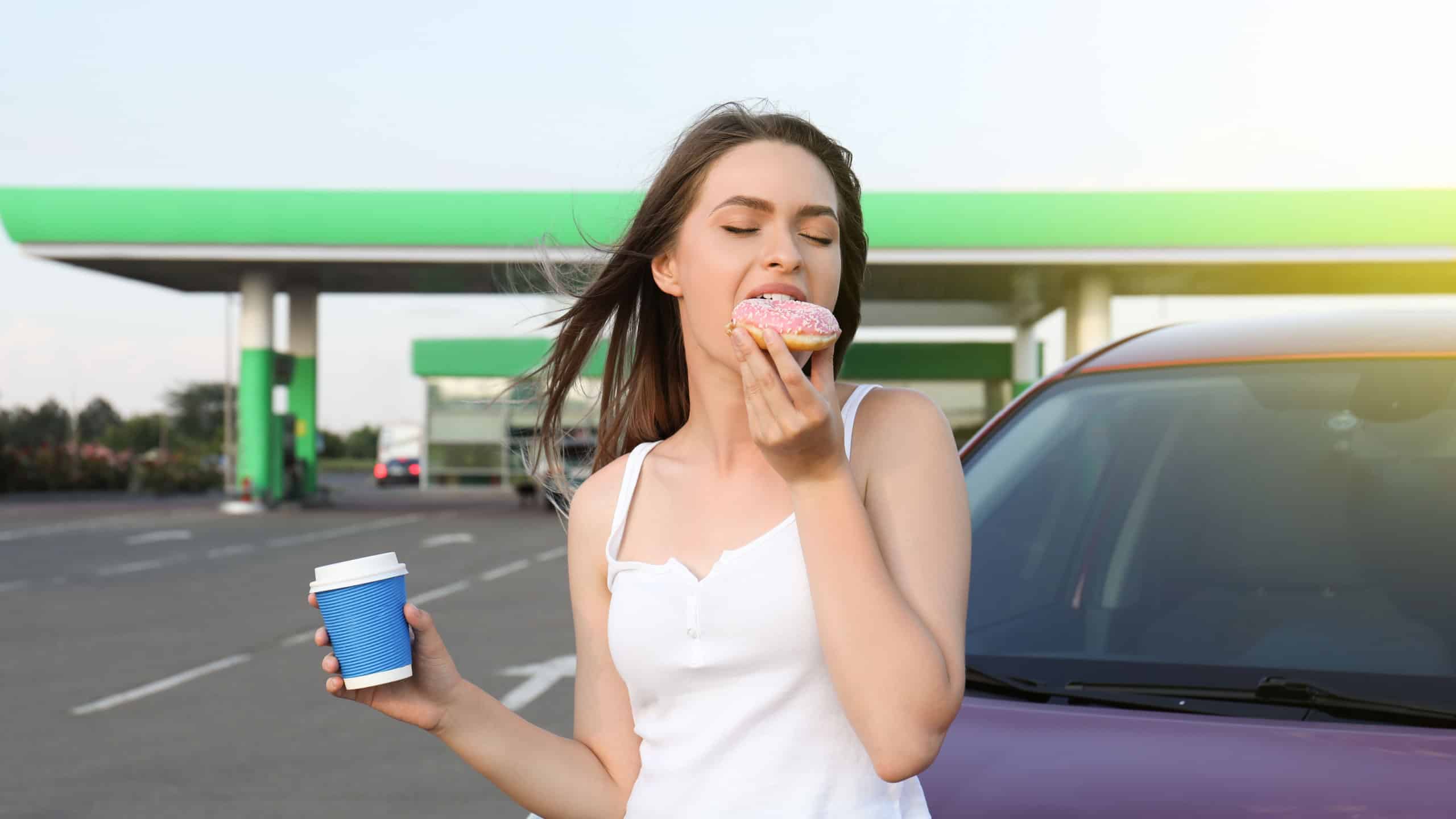 Beautiful young woman with coffee eating doughnut near car at gas station