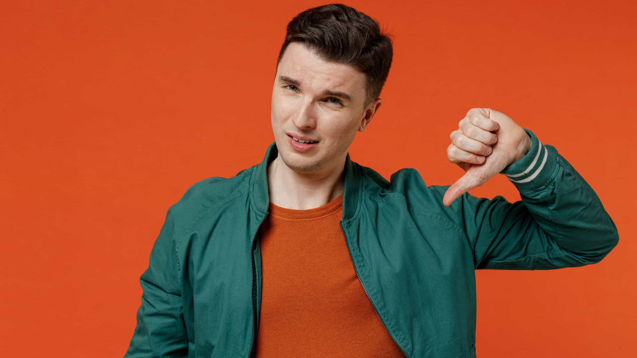 Sceptical frowning cynical young brunet man 20s wears red t-shirt green jacket showing thumb down dislike gesture isolated on plain orange background studio portrait. People emotions lifestyle concept