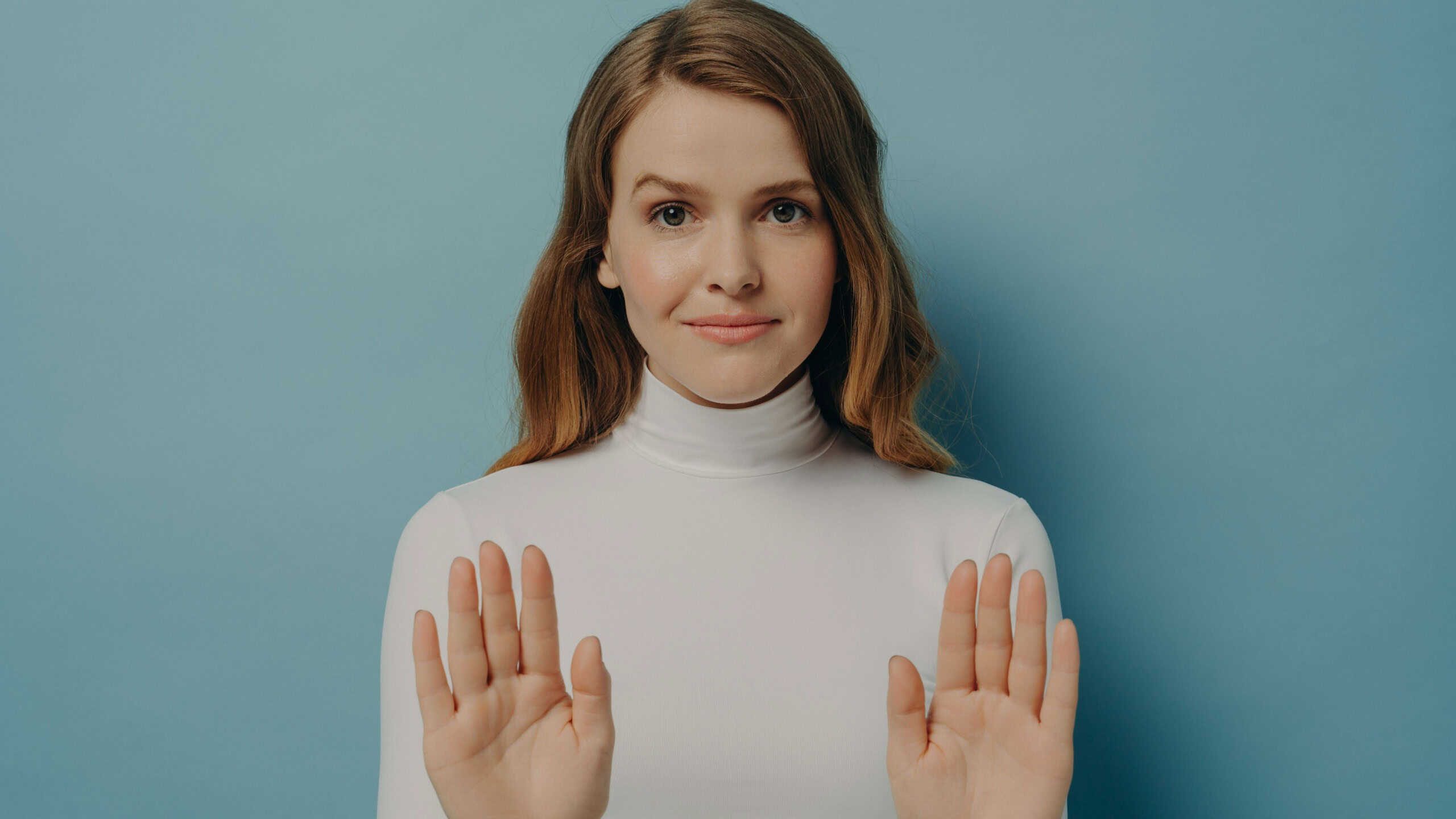 Confident young woman showing her palms while keeping both hands outstretched in stop gesture isolated over blue studio wall background, caucasian female saying no. Refusement concept