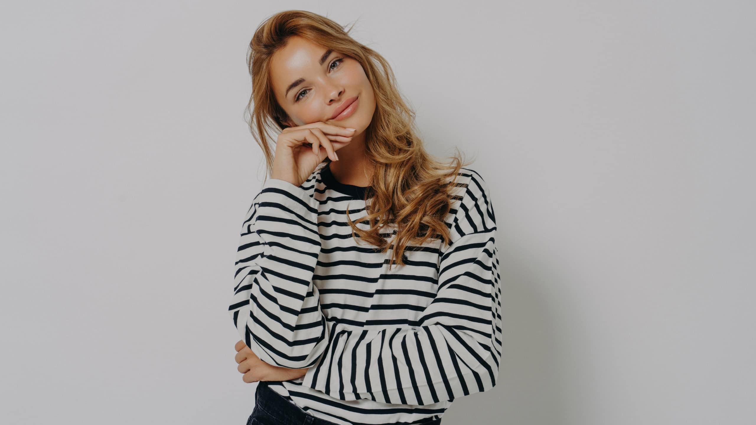 Studio shot of satisfied young European woman keeps hand on face tilts head looks gently at camera weras casual striped jumper and jeans isolated over grey background. Human face expression concept