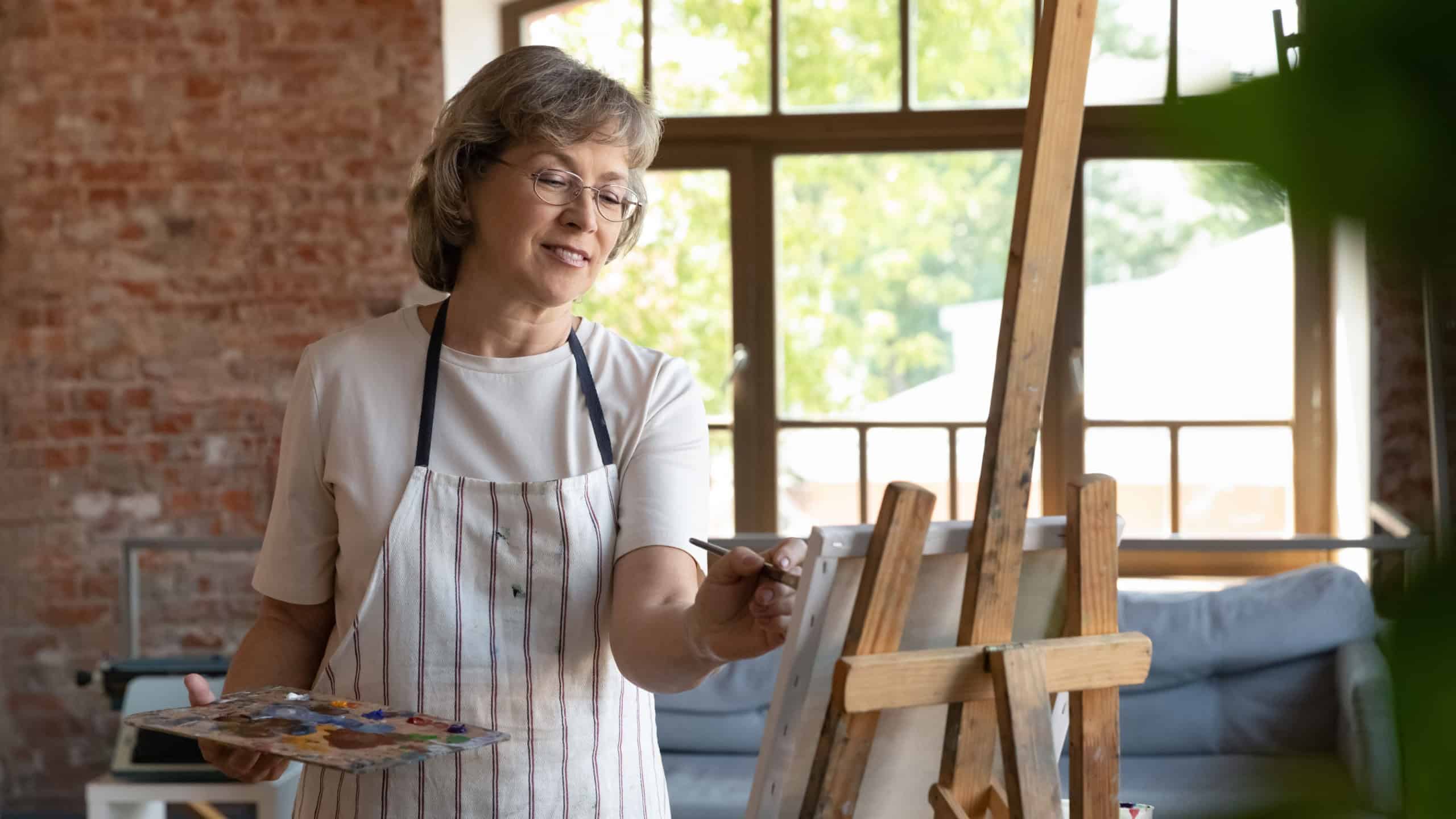 Happy senior retired lady drawing in paints on canvas. Elder talented professional artist working at easel in artistic studio, using palette, paintbrush, smiling, enjoying creativity, inspiration