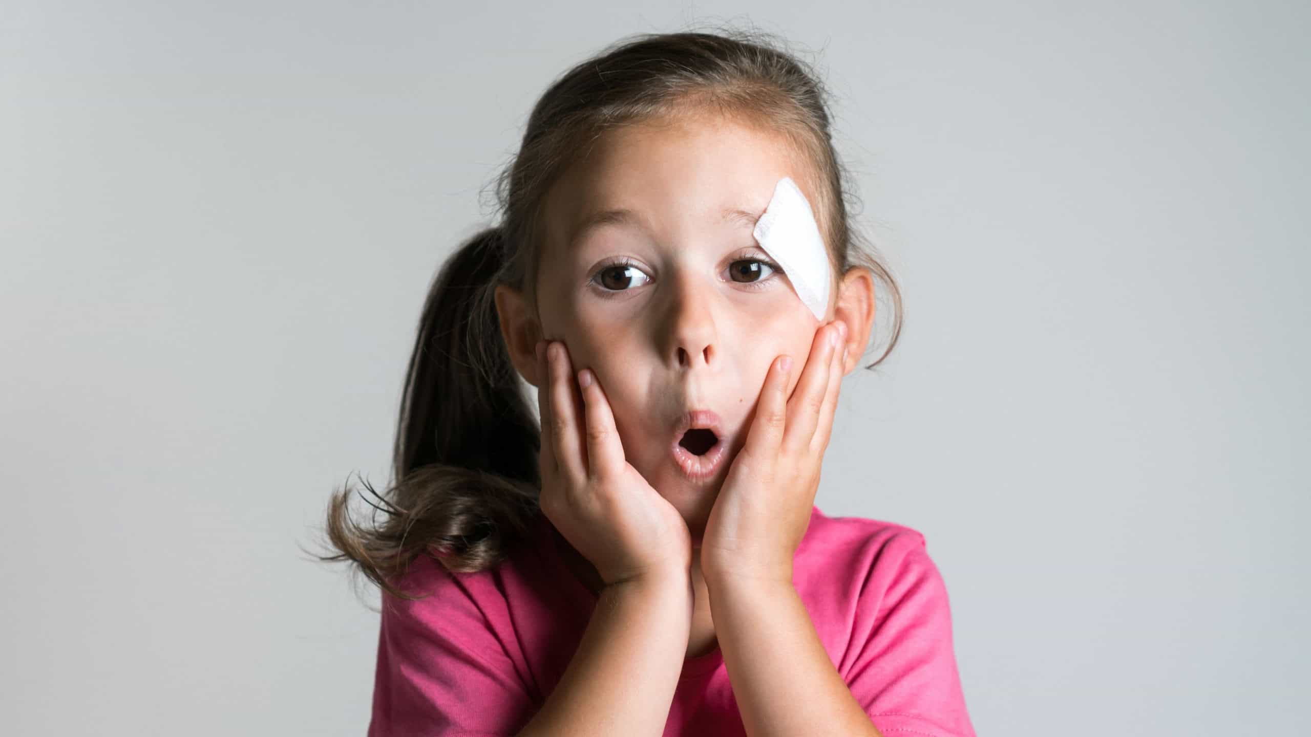 Close up surprised, cute, nice dark haired little girl with bandage patch on face, wear pink shirt on white background. Afraid of parents reaction. Physical wounds, fidget children, doctor fear