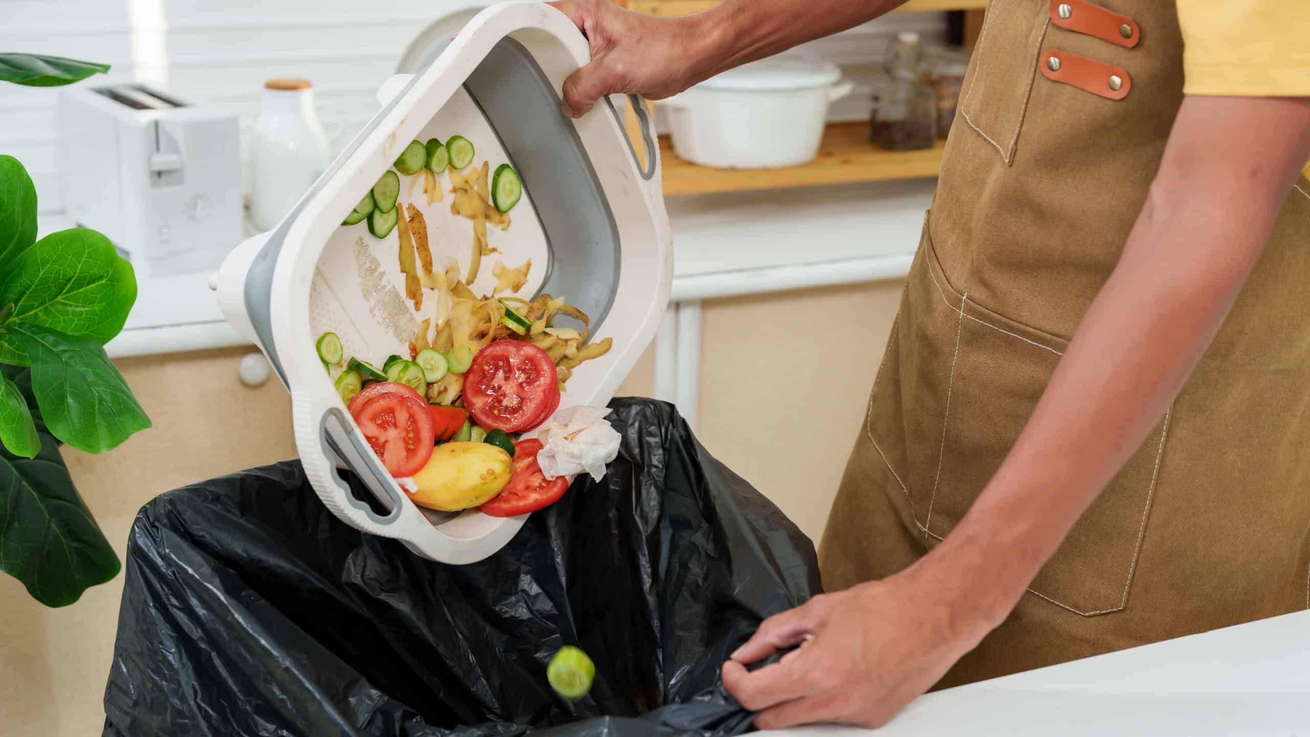 Close-up shot of a man's hand carrying food scraps and unused vegetable scraps Throw it in the trash with black bags. to separate different types of waste Wearing an apron in kitchen. stay home alone