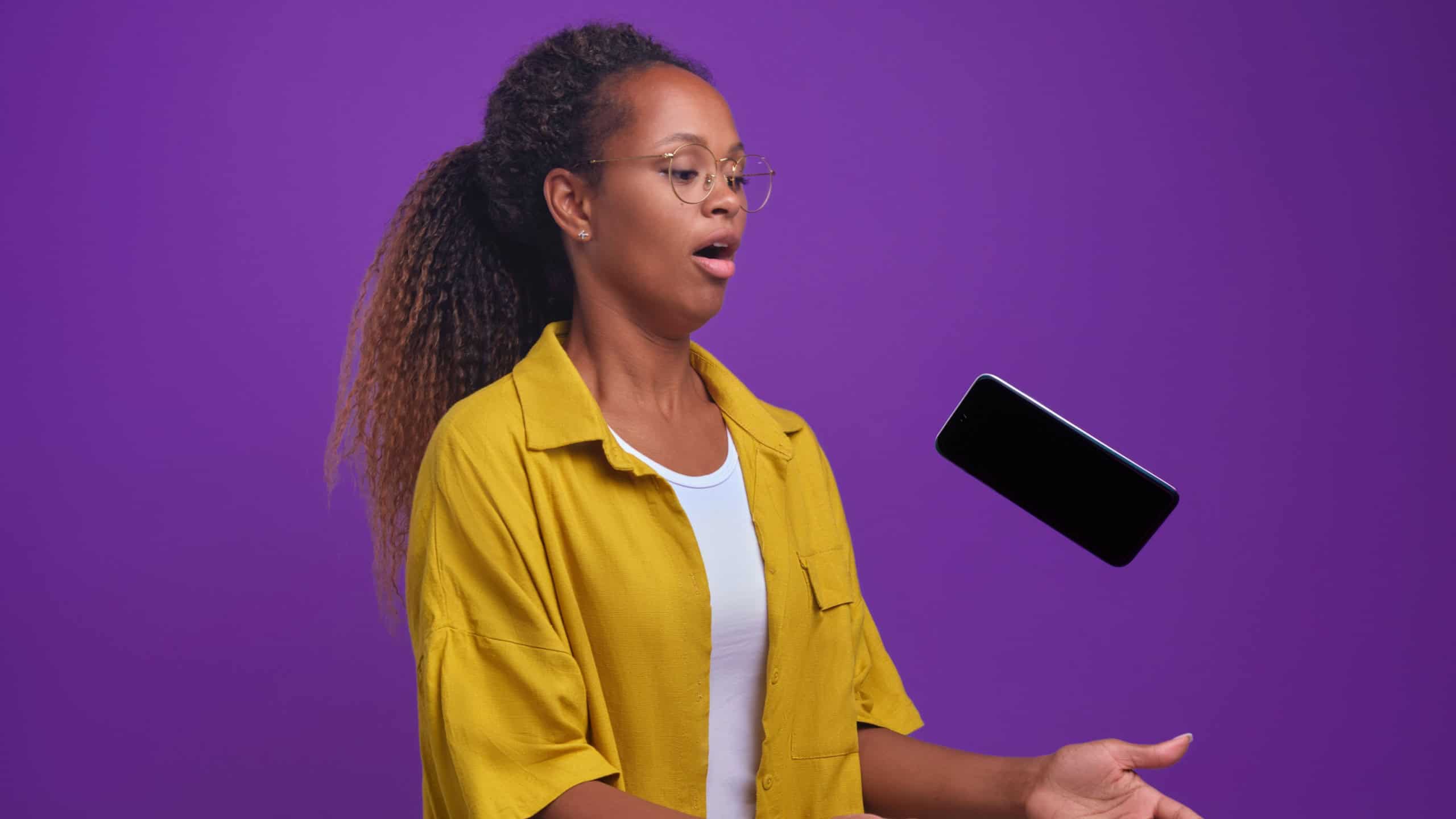 Young scared African American woman trying to catch falling mobile phone suffering from clumsiness or coordination problems causing costly gadgets to be dropped posing on isolated lilac background