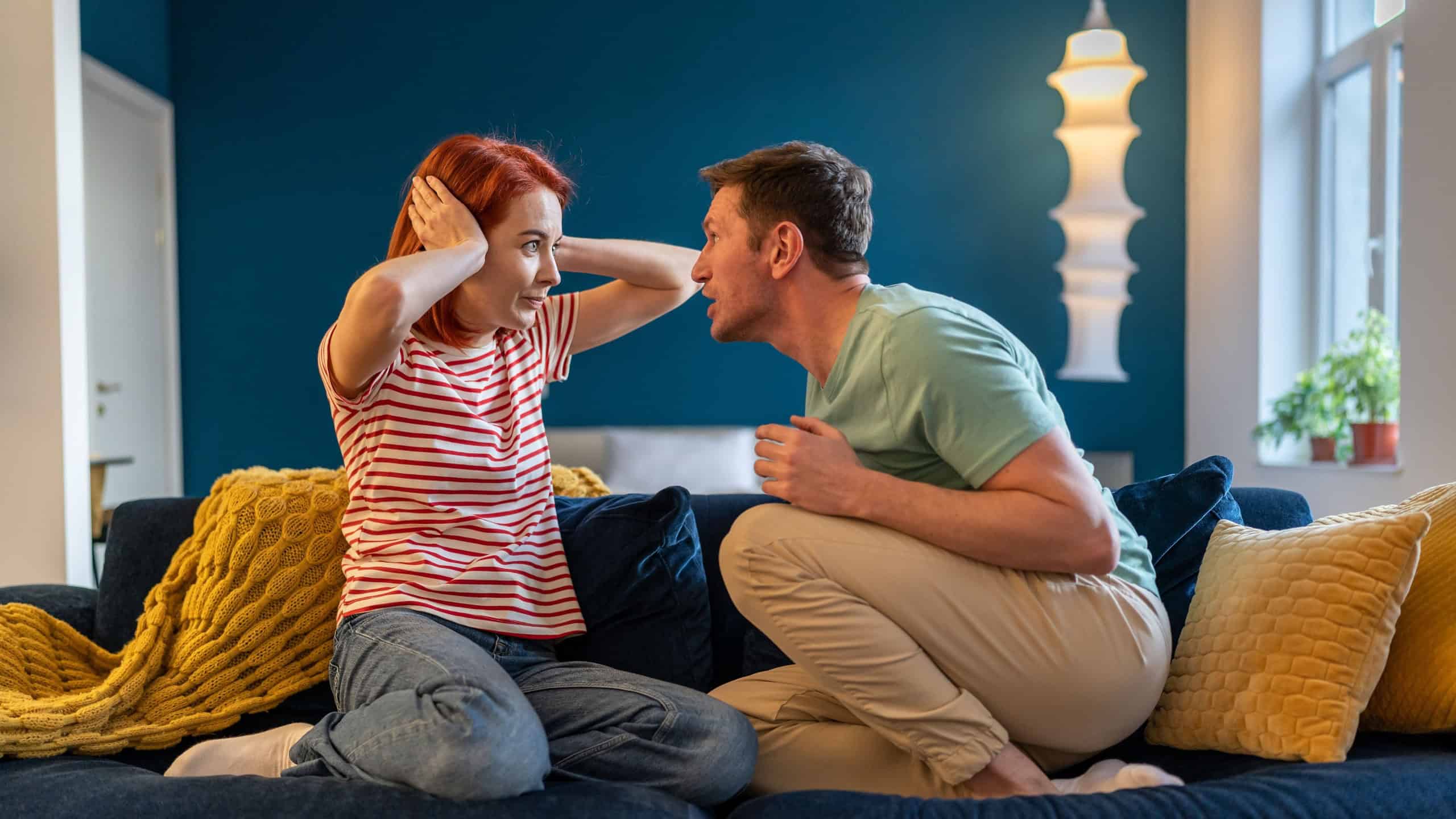 Tyrant husband shouting on wife sitting on couch at home looking at her face. Scared woman closing ears. Domestic violence, abuse, neurotic relationships, toxic couple relations, marital discord.