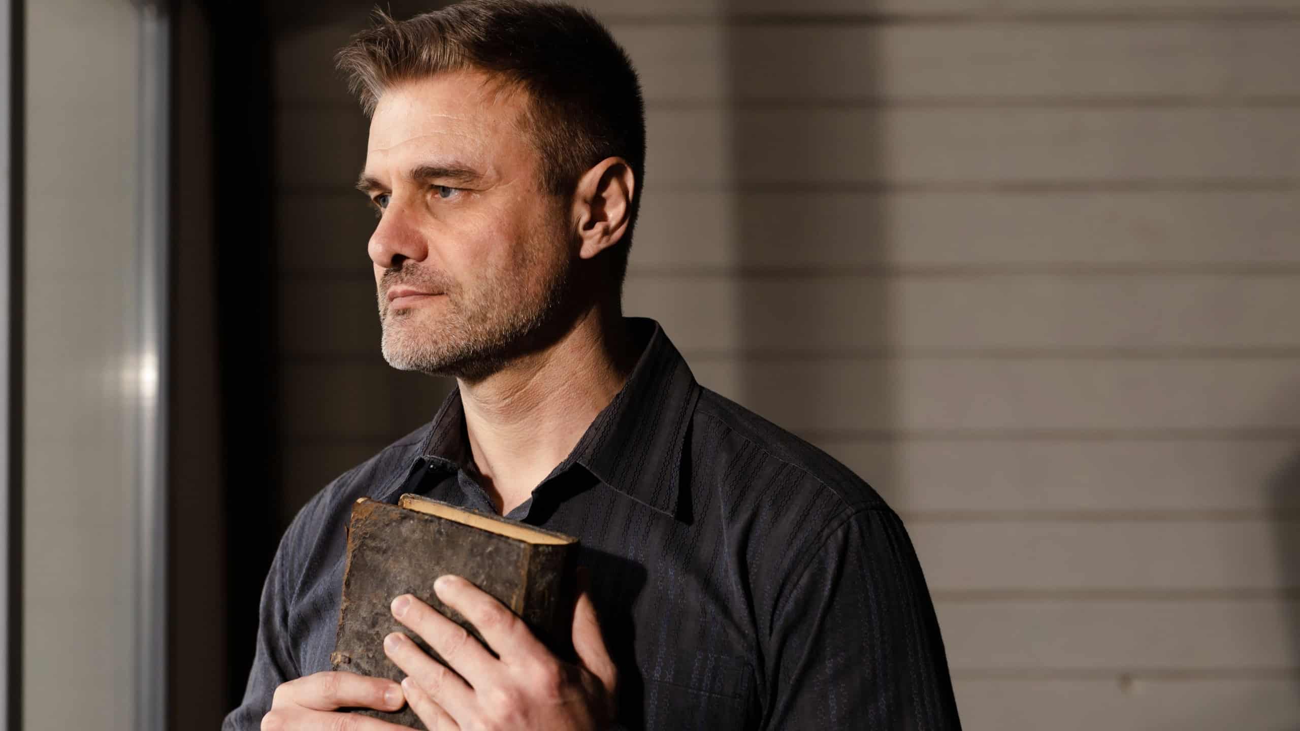 Young caucasian confident man in black shirt hugging bible to himself. Male praying god with hands on great holy book indoors. Christian adult guy holding scripture