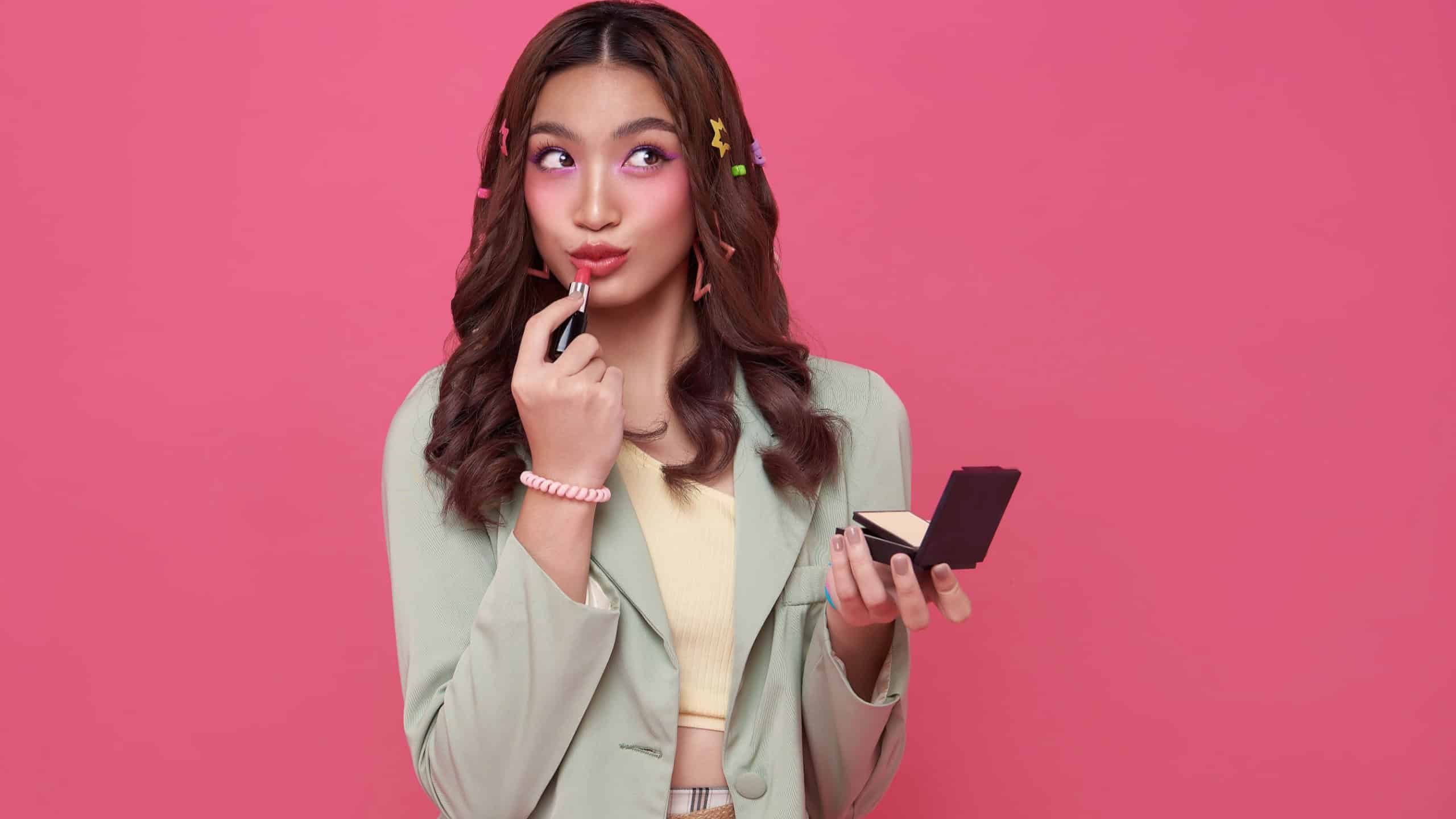 Makeup lipstick woman ready looking in pocket mirror. beautiful trendy young asian female fashion model putting makeup on pink background.