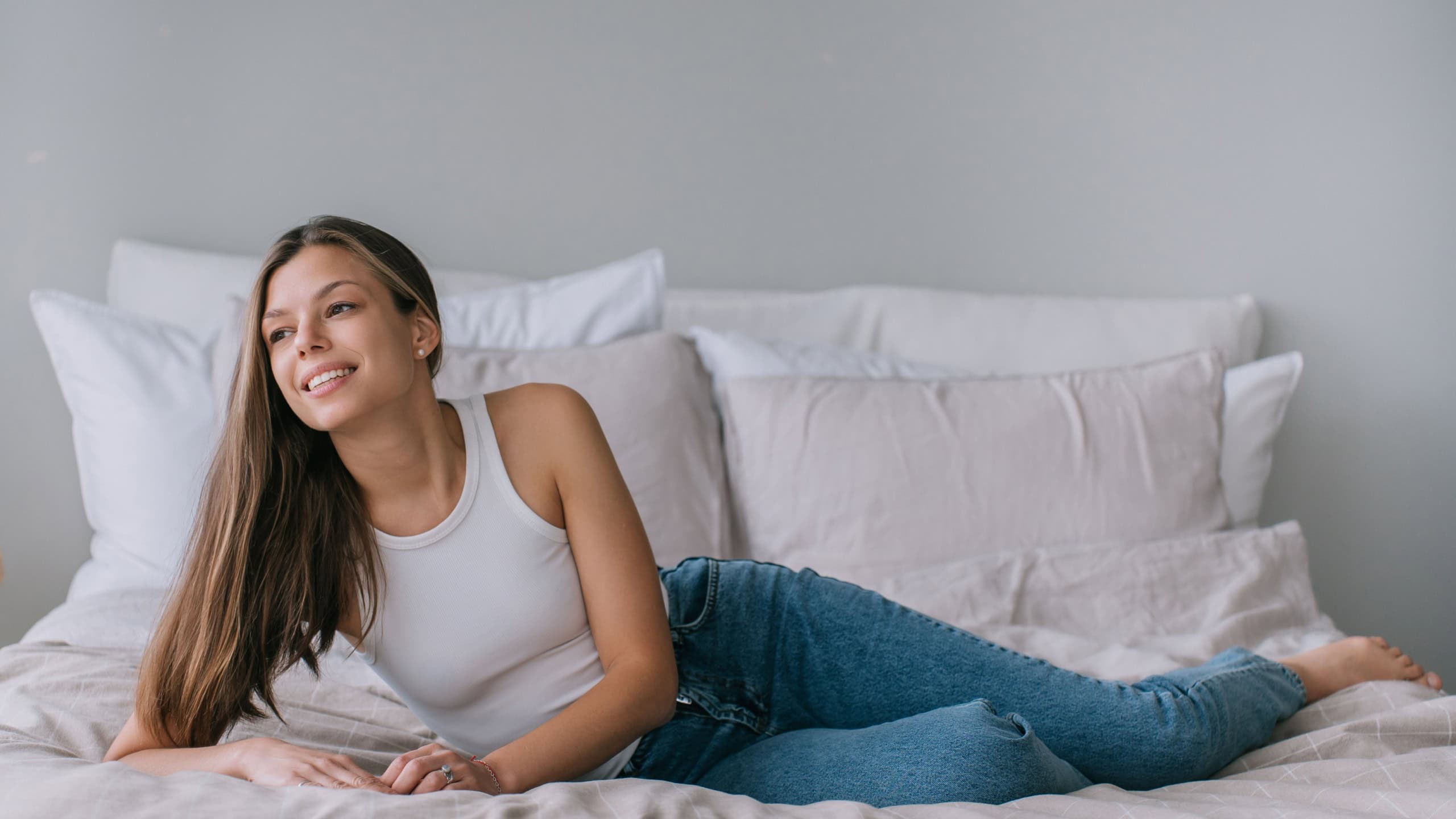 Relaxed young woman lounging on a bed, casually dressed in a white tank top and blue jeans, with a serene smile, embodying comfort and effortless style in a cozy setting.