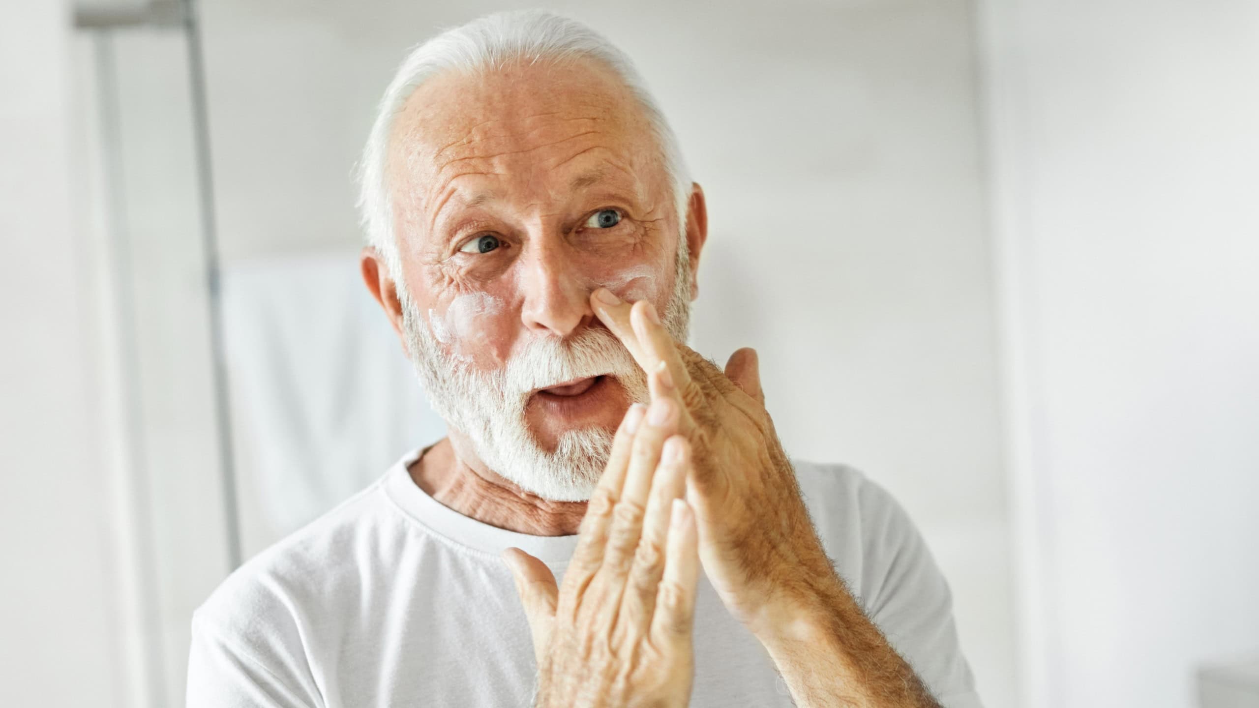 Portrait of an elderly senior man applaying a cream on his face in front of a mirror in bathroom, skincare, beauty and vitality concepts