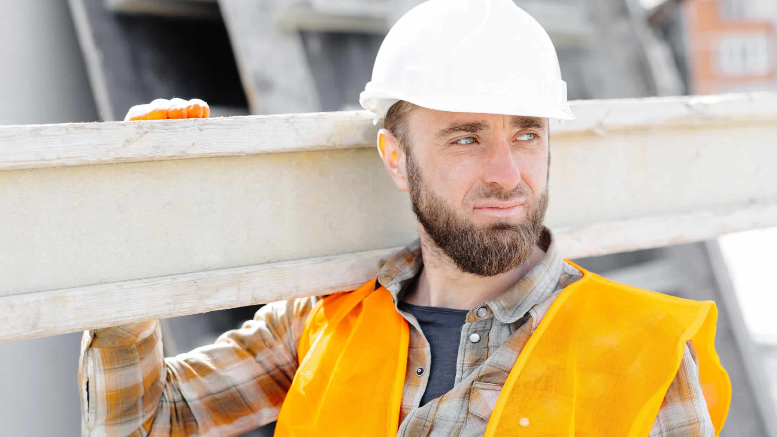 Builder man in hardhat and vest carrying timber on building site in his working day.