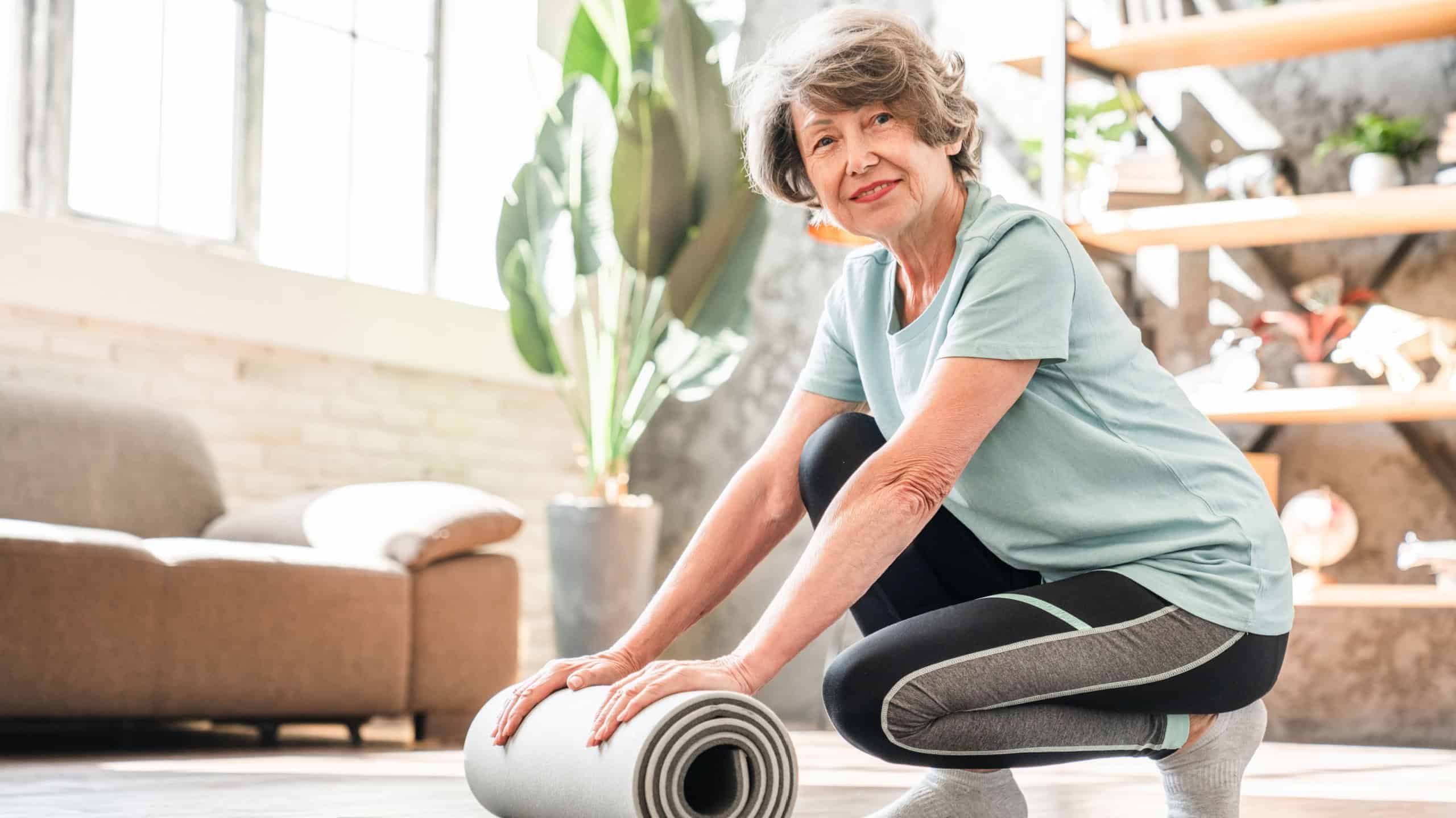 Fit sporty old senior woman rolling up the fitness mat after training at home. Body shaping, slimming physical exercises for active seniors