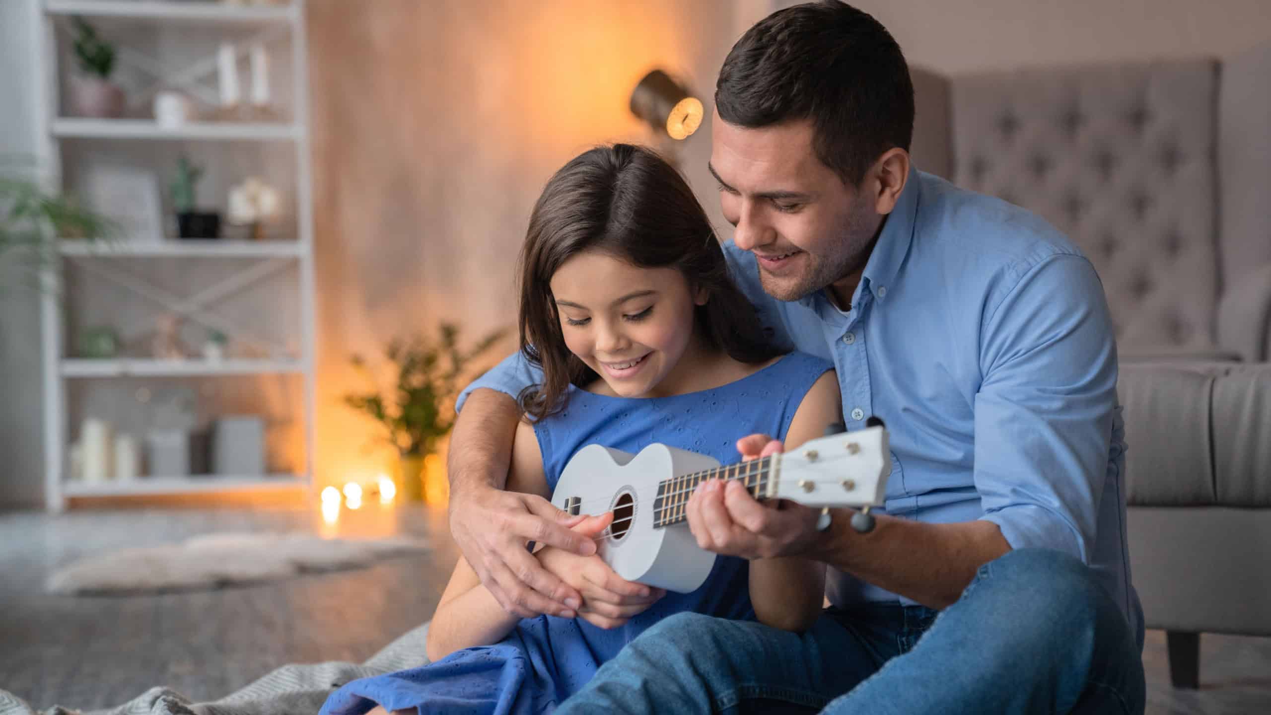 Cute little smiling girl playing on white wooden ukulele guitar with her smiling cheerful daddy dad father. Happy father teaching to play on ukulele guitar his daughter at cozy home