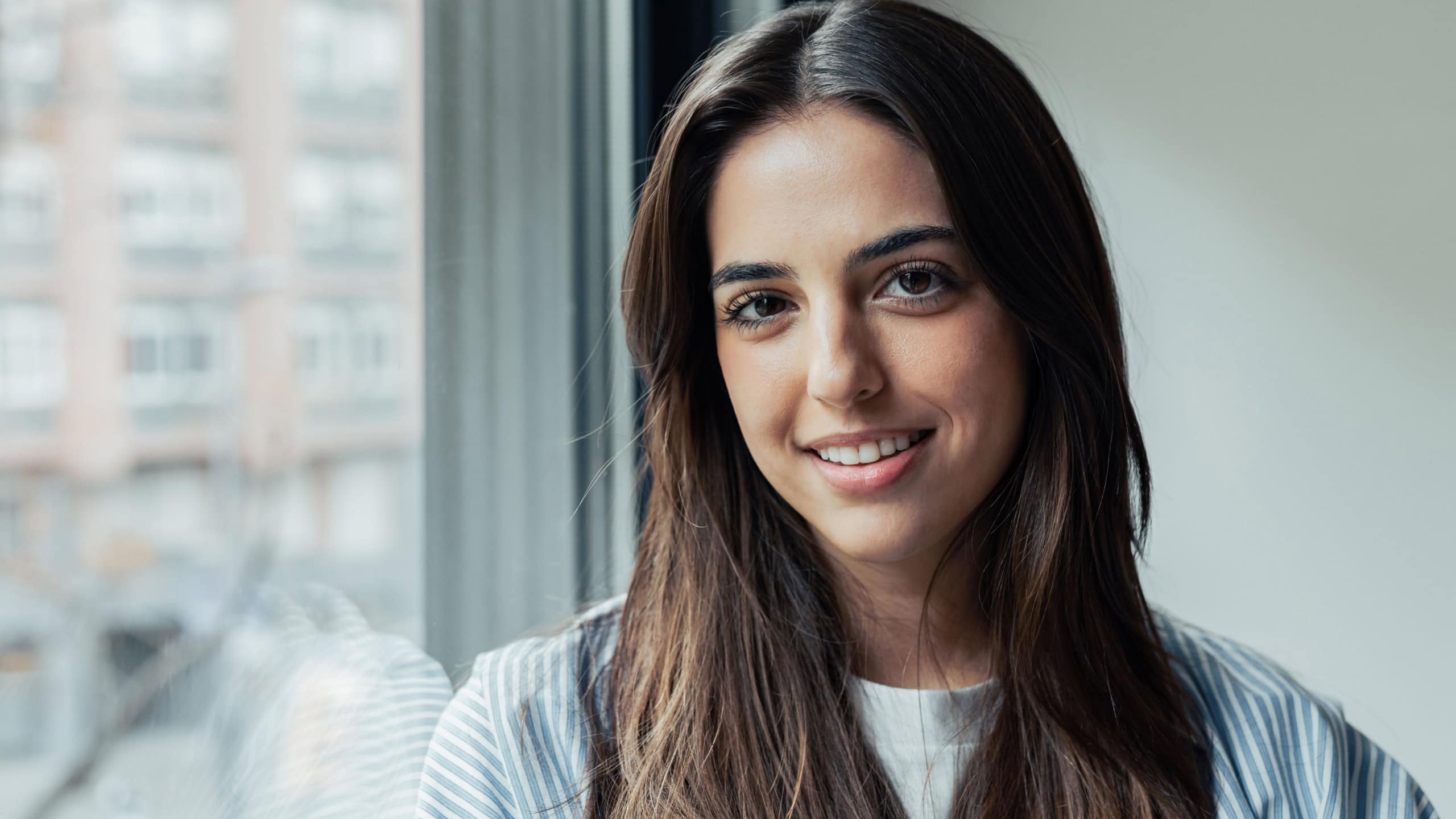 Head shot portrait confident positive woman with healthy toothy smile looking at camera, standing near window, successful happy businesswoman entrepreneur student posing for profile picture