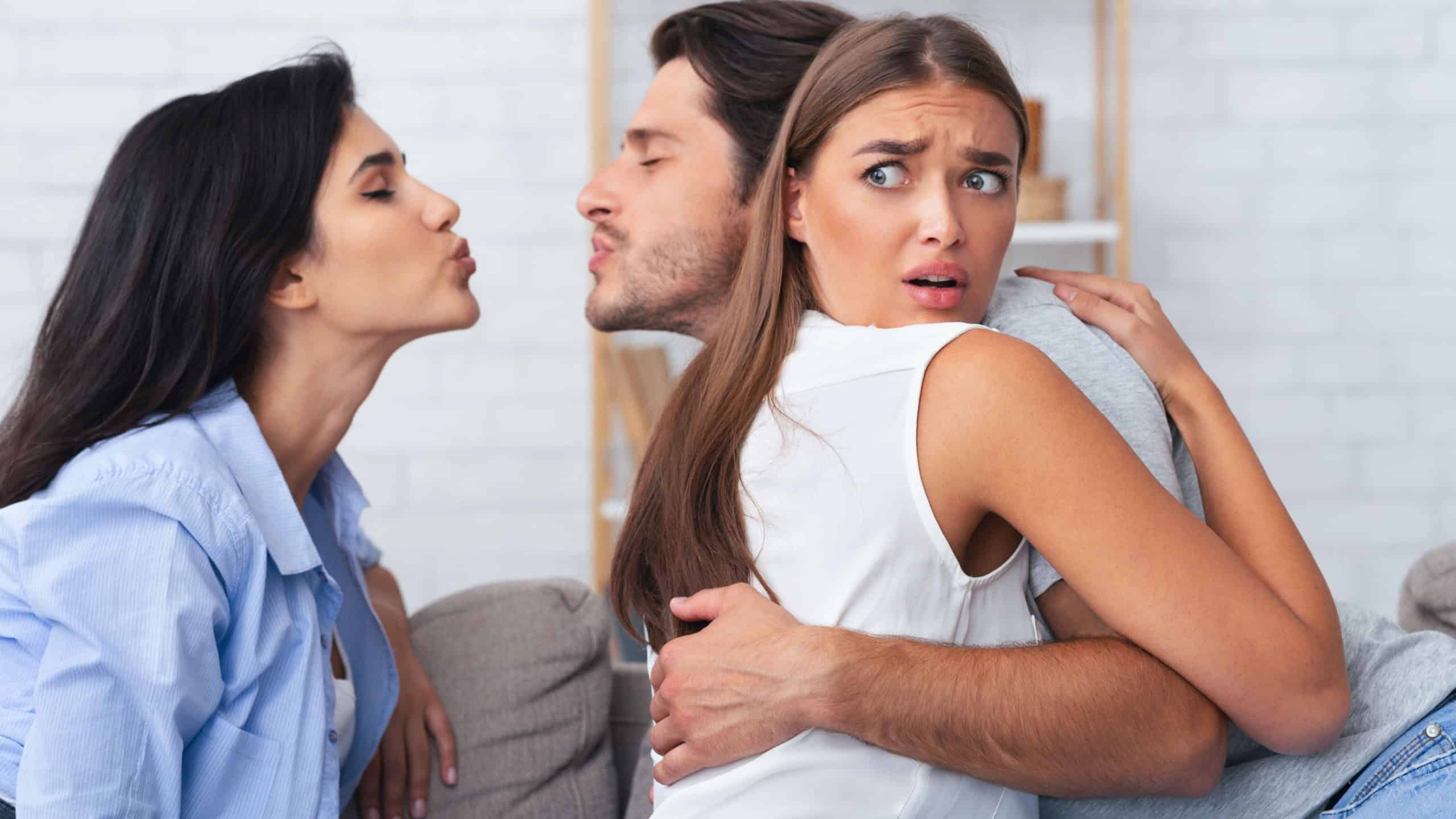 Jealousy And Infidelity. Girl Hugging Cheating Boyfriend While He Kisses Her Friend Sitting Together On Sofa At Home
