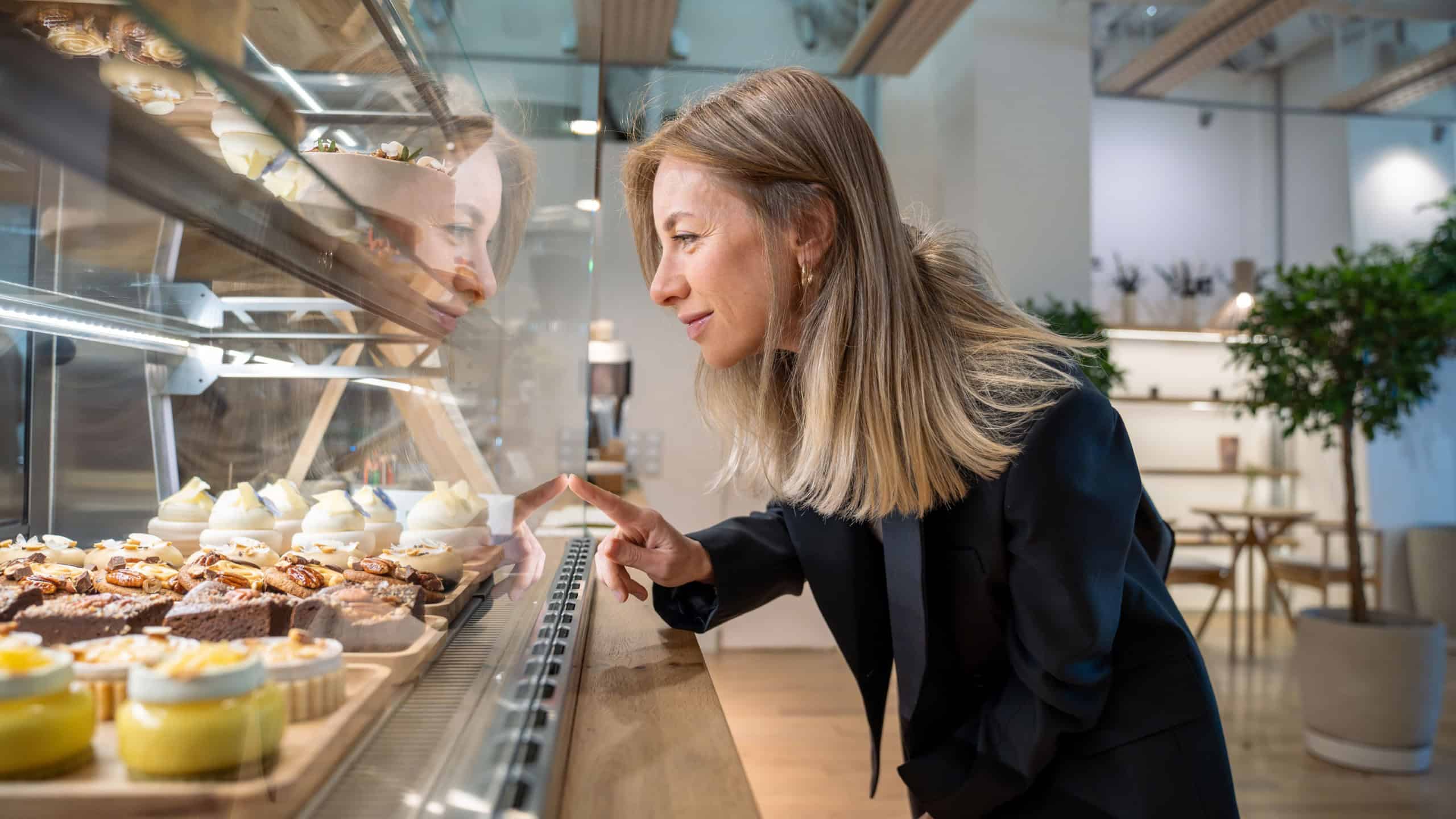 Happy woman looking at snacks for coffee, standing at showcase in cozy small cafe, going to buy purchase lunch, breakfast. Satisfied cheerful businesswoman at window display in coffeehouse, bakehouse.