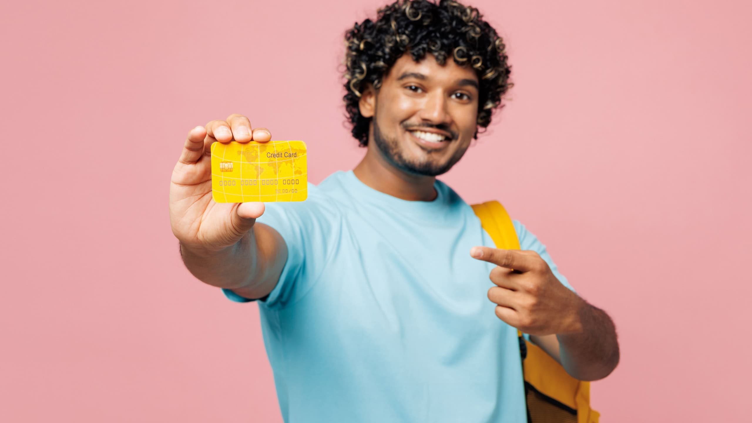 Young smiling smart Indian boy student wear blue t-shirt casual clothes backpack bag hold point on close up credit bank card isolated on plain pink background. High school university college concept
