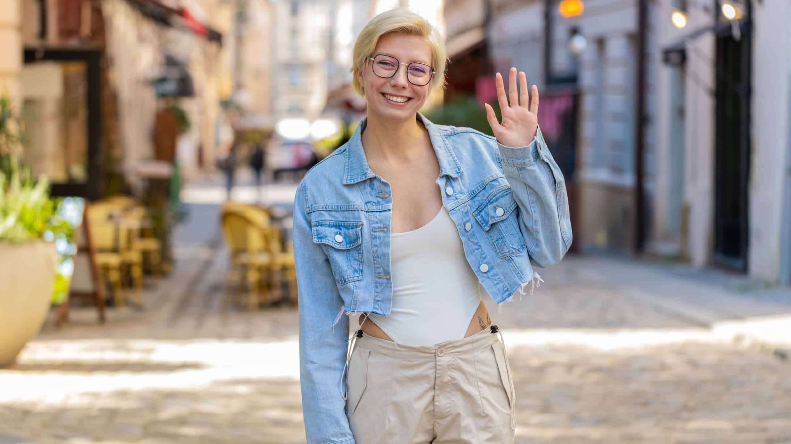 Cheerful young woman tourist smiling friendly at camera, waving hands gesturing invitation hello, hi, greeting, goodbye, welcoming with hospitable expression outdoors. Lady in glasses on city street.