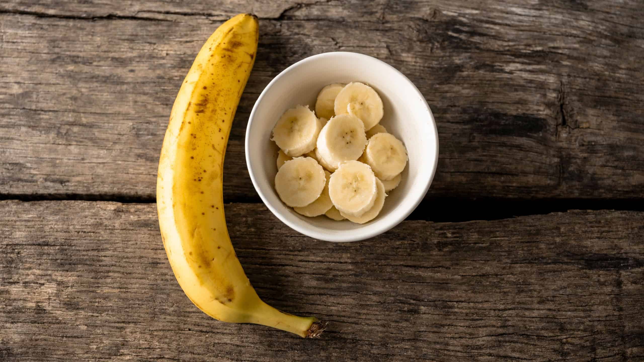 Top view of fresh banana slices in a cup next to a whole ripe banana placed on rustic wooden boards.
