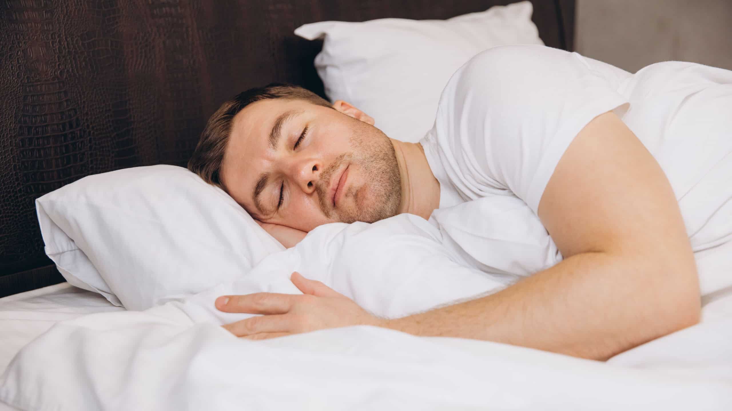 Young man sleeping peacefully in bed enjoying good sleep, resting on white soft pillows, covered with blanket, healthy lifestyle concept