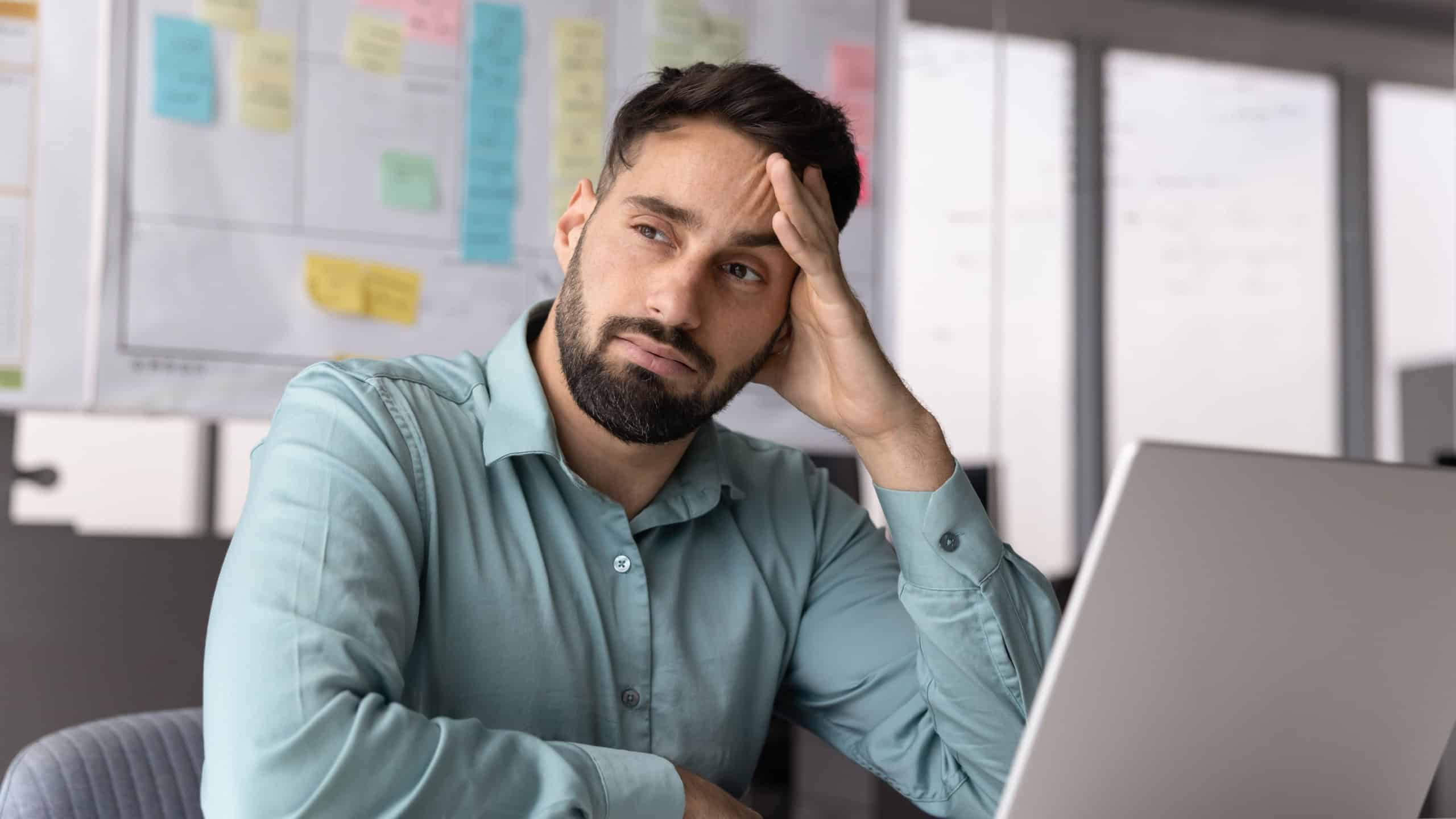 Thoughtful tired Hispanic businessman in casual thinking on project solutions, doubts at laptop, making decision, leaning head on hand, looking away, sitting at work table with laptop