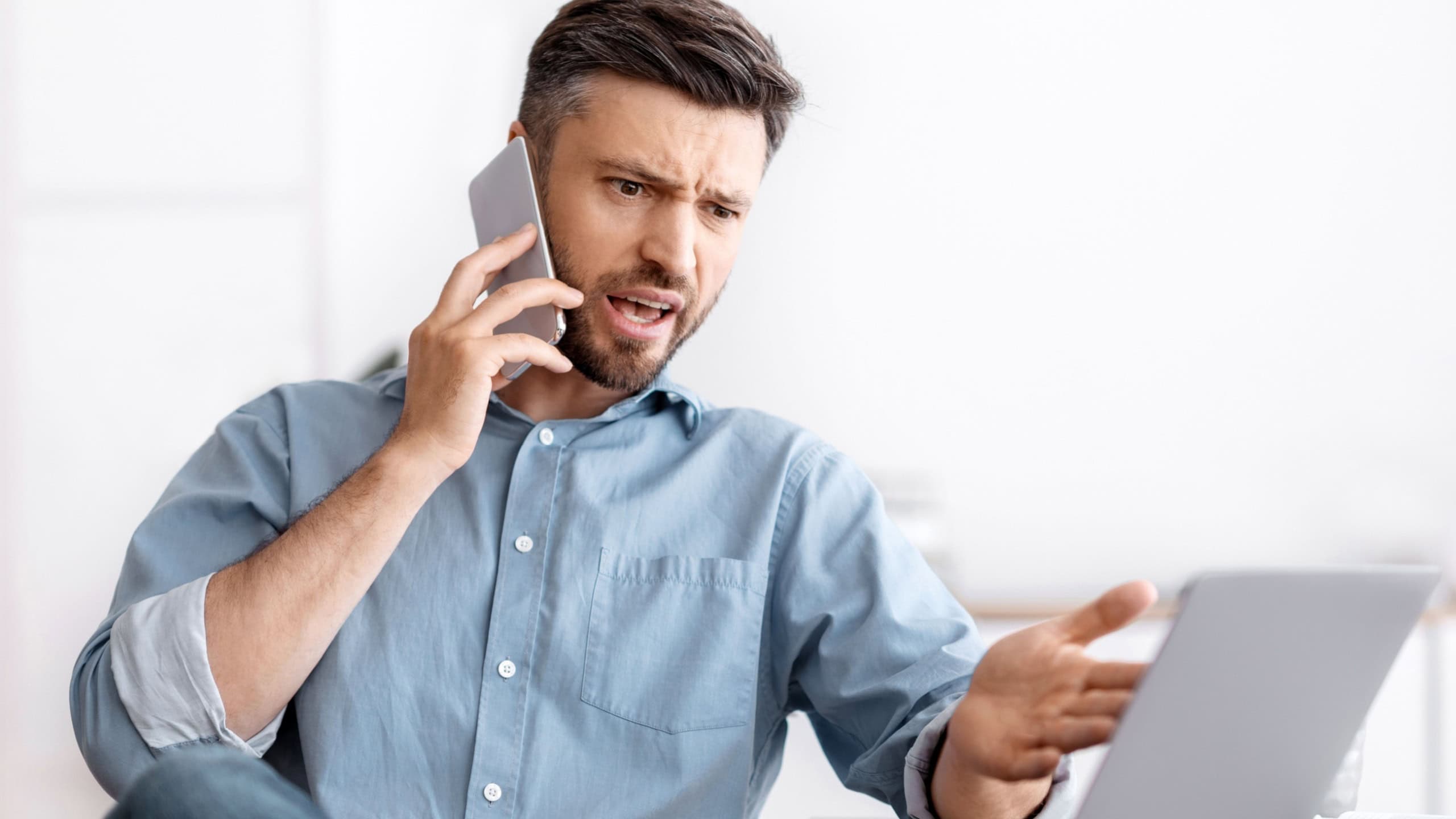 Angry millennial businessman talking on mobile phone and looking at laptop screen working in modern office, having problems with project, annoyed boss speaking with employee by phone, copy space