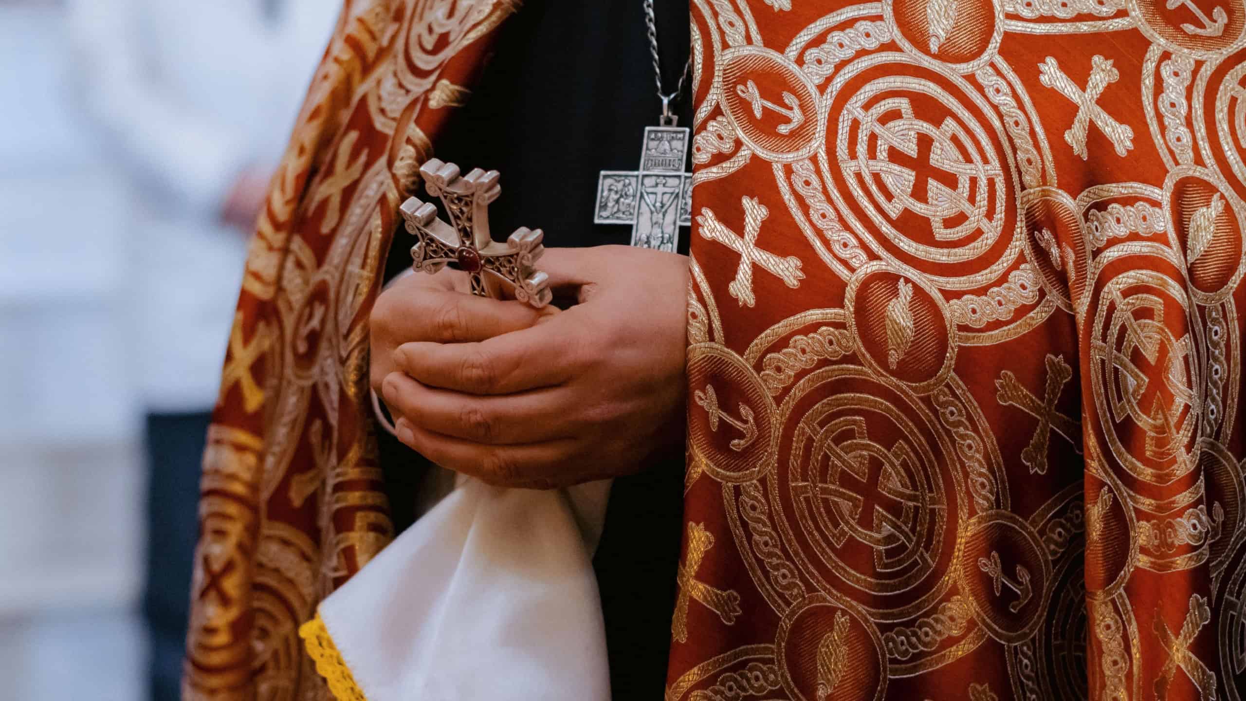 A priest's hands holding a cross and cloth, wearing ornate vestments during a religious ceremony.