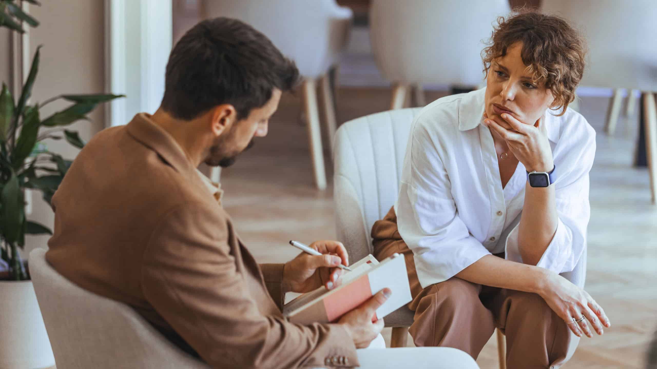A counselor and a client are seated in a well-lit room, sharing a serious yet calm dialogue. The image portrays a professional setting focusing on therapy and support.