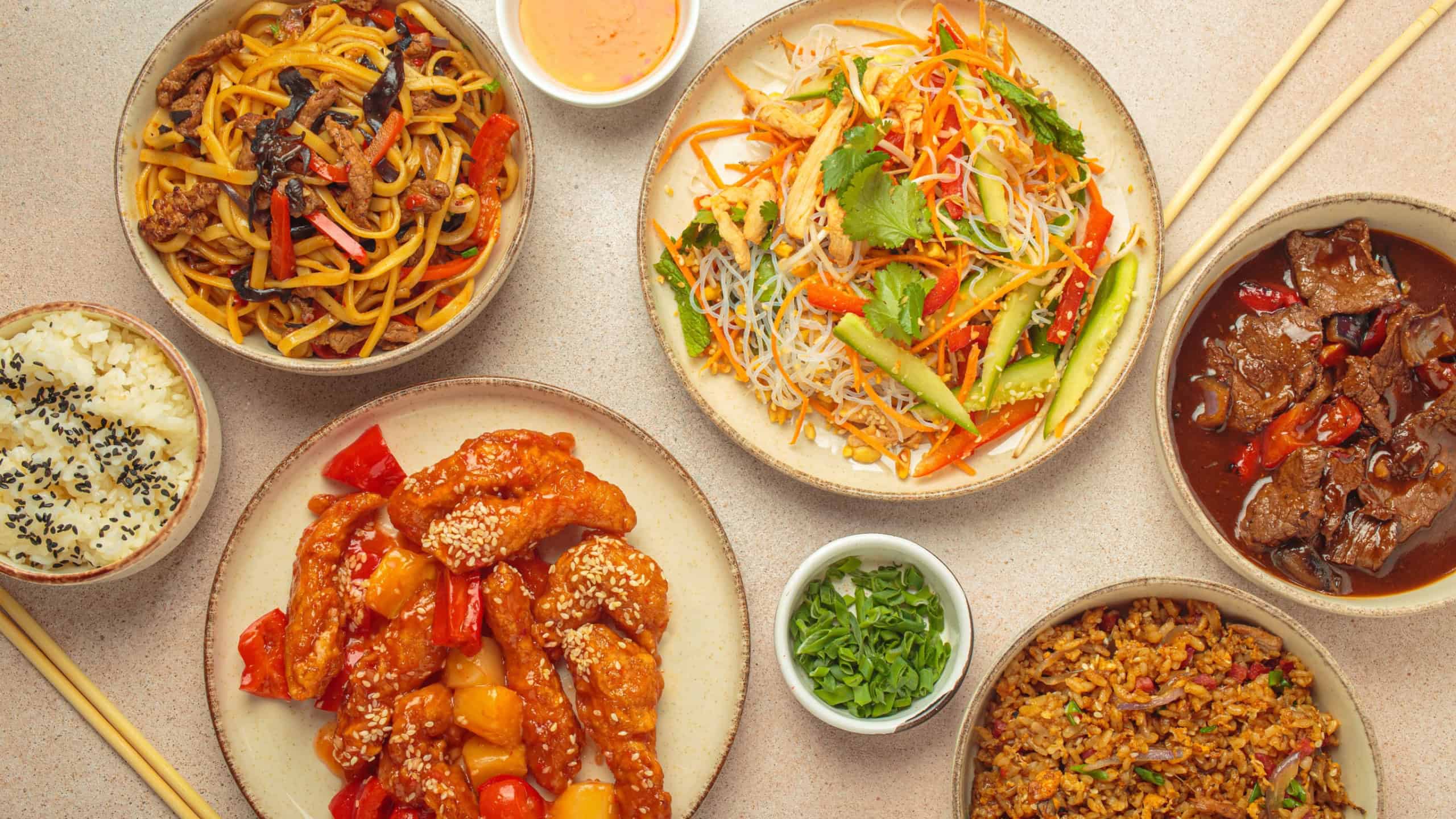 Variety of Chinese food dishes top view on table with chopsticks, light beige background, cuisine of China