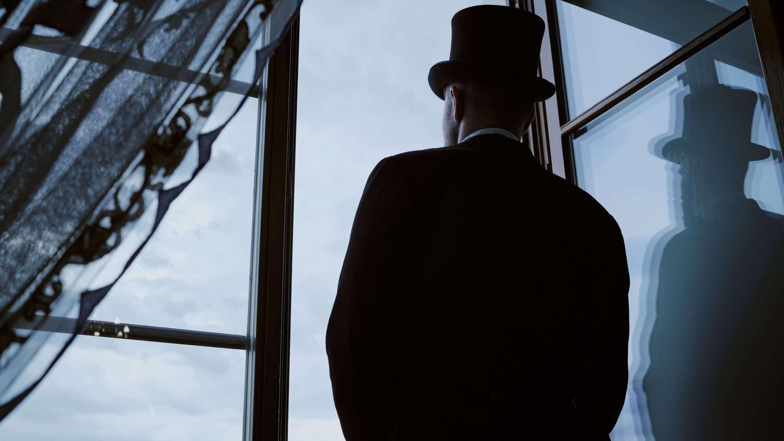 Caucasian middle aged man wearing top hat standing by window gazing outside, partial silhouette visible, facial features obscured, Victorian style suggested by attire and posture