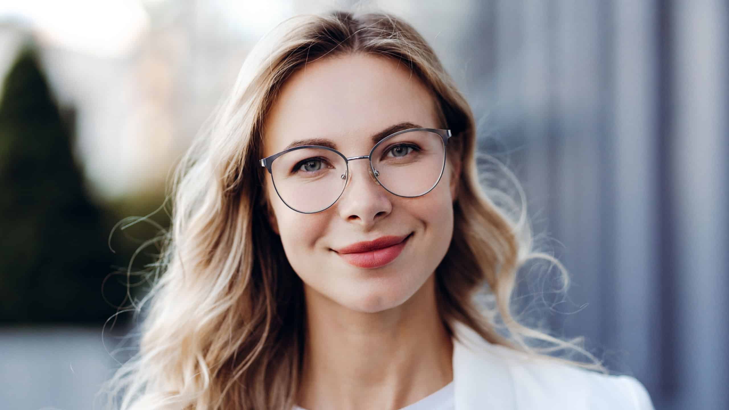 Blonde professional woman with glasses smiling confidently in natural outdoor light, headshot conveying success and leadership