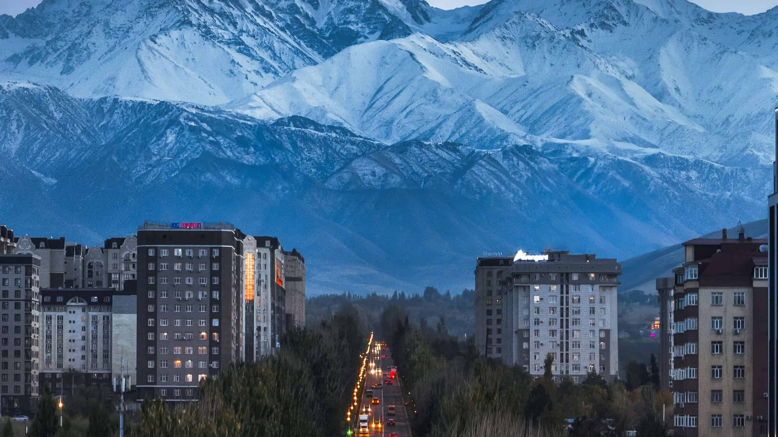 Bishkek, Kyrgyzstan - May 12, 2024: Evening view of a city boulevard with car taillights tracing lines, framed by illuminated apartment buildings and snow-covered mountains