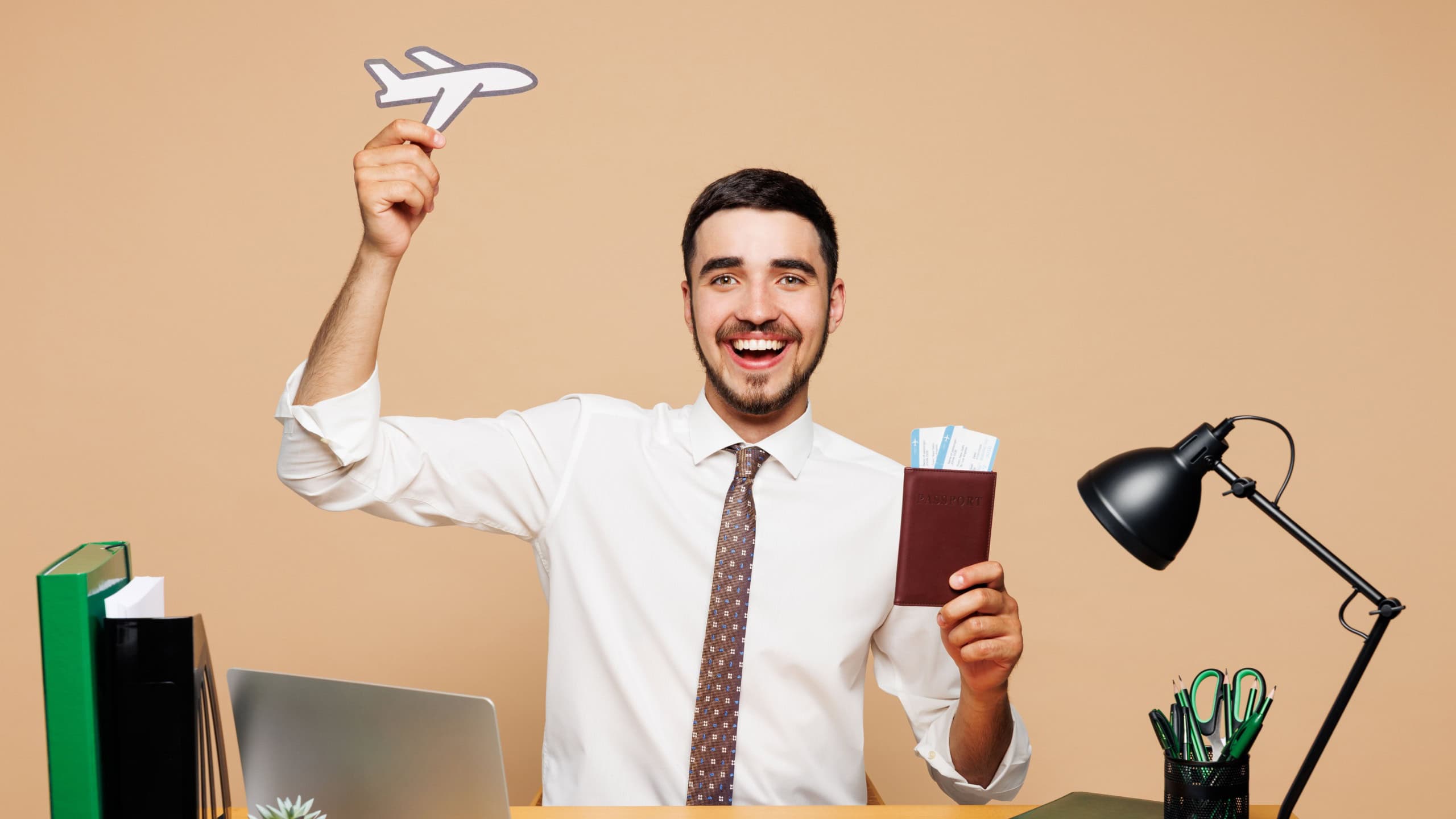 Young traveler tourist employee business man wear white shirt sit work at office desk travel abroad hold passport ticket airplane isolated on plain beige background. Air flight trip journey concept