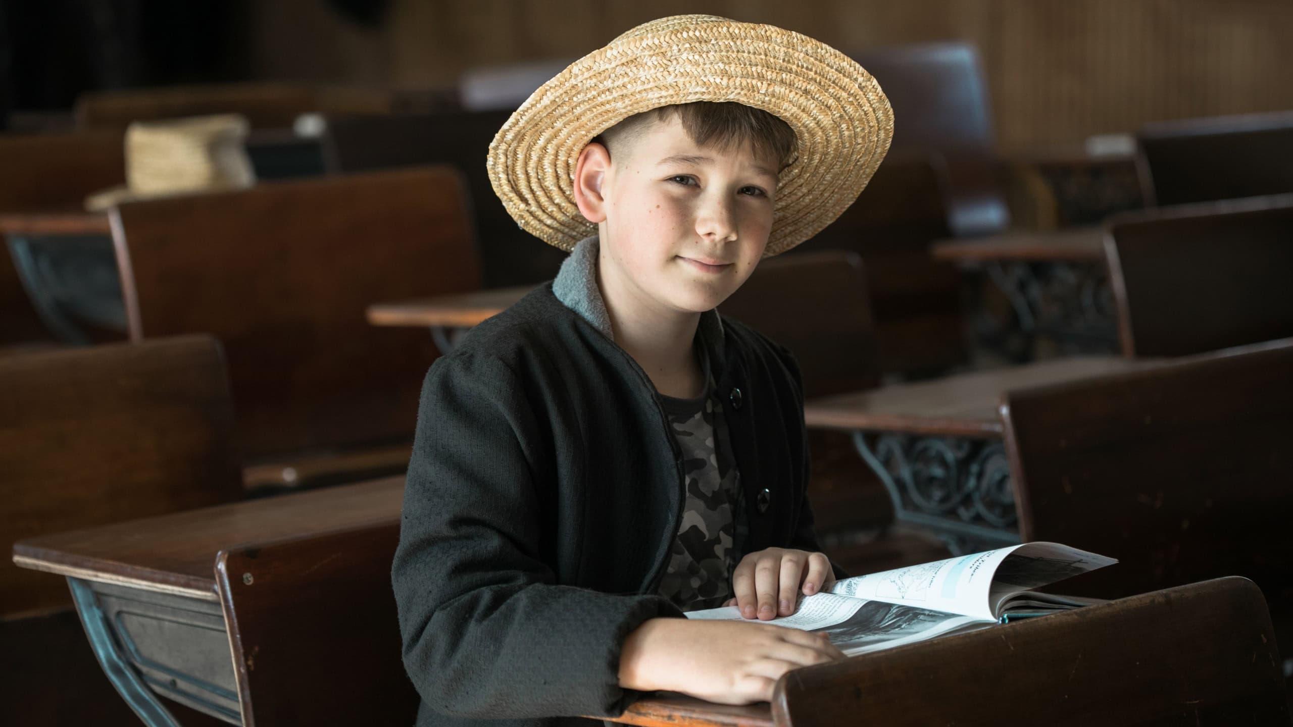 Amish boy sitting at an old wooden school desk with a book in vintage classroom.