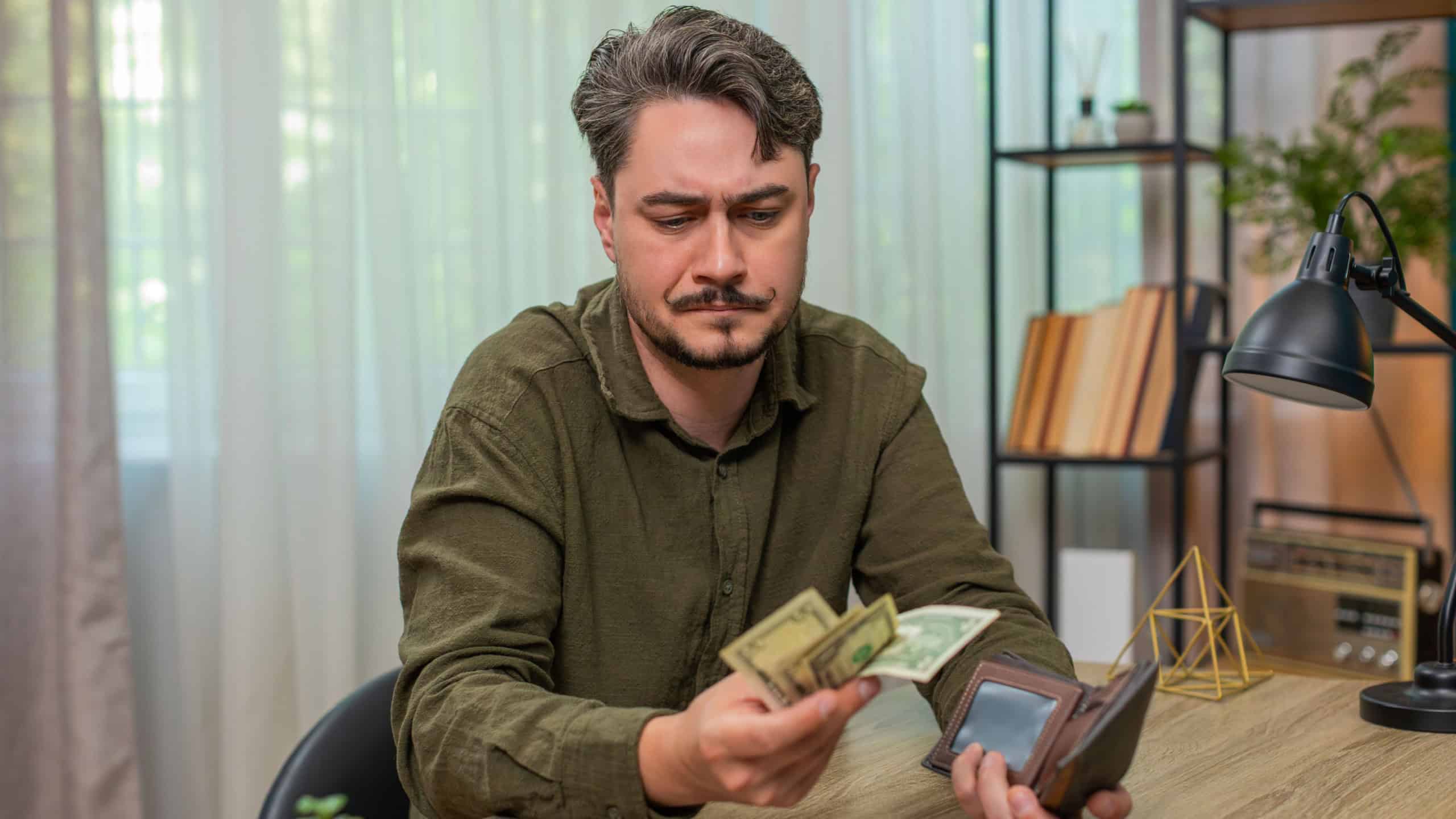 Poor sad man at home counts few cash bills frowns shaking head worried about debt poverty and money shortage. Young adult guy at table sorts dollars feeling stress fear bankruptcy serious trouble