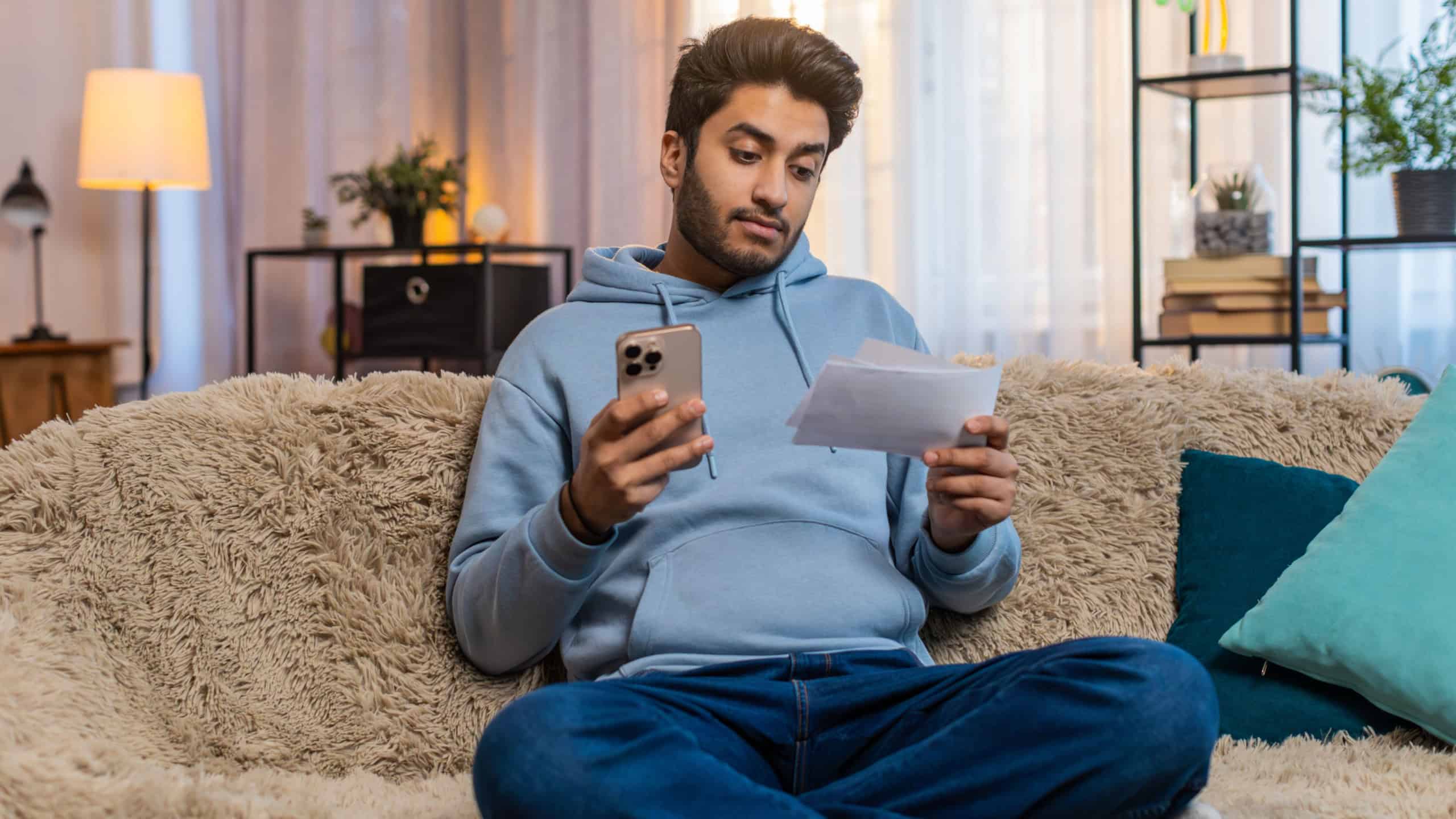 Young Indian man paying utility bills through app holding smartphone and receipts sitting on sofa in living room at home. Hispanic guy reviews amounts attentively ensuring accurate payment details.