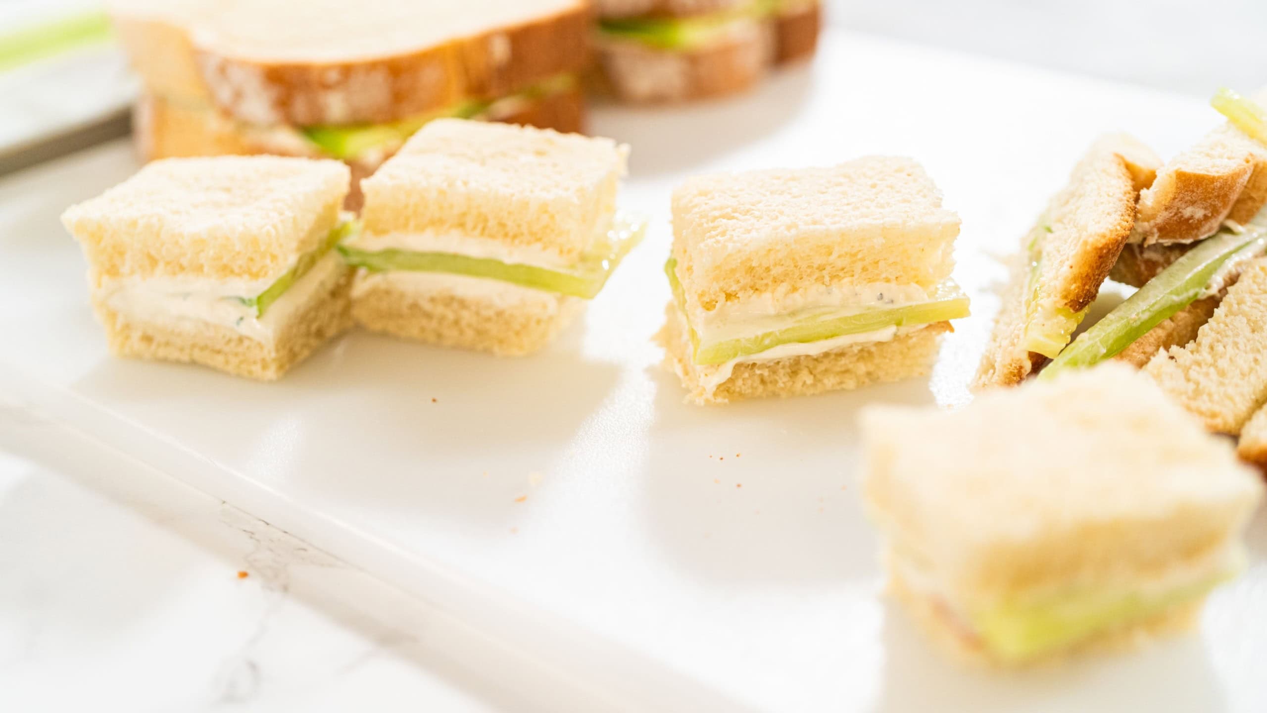 Small square cucumber tea sandwiches with white bread and cream cheese are neatly placed on a white cutting board. The sandwiches are sliced evenly and arranged in rows.