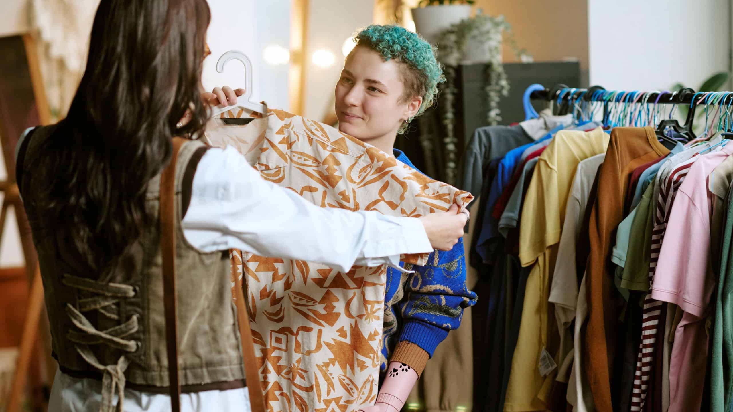 Young Caucasian woman with short curly hair and prosthetic arm assisting another young woman choosing second hand clothing in thrift store, both standing near clothing rack