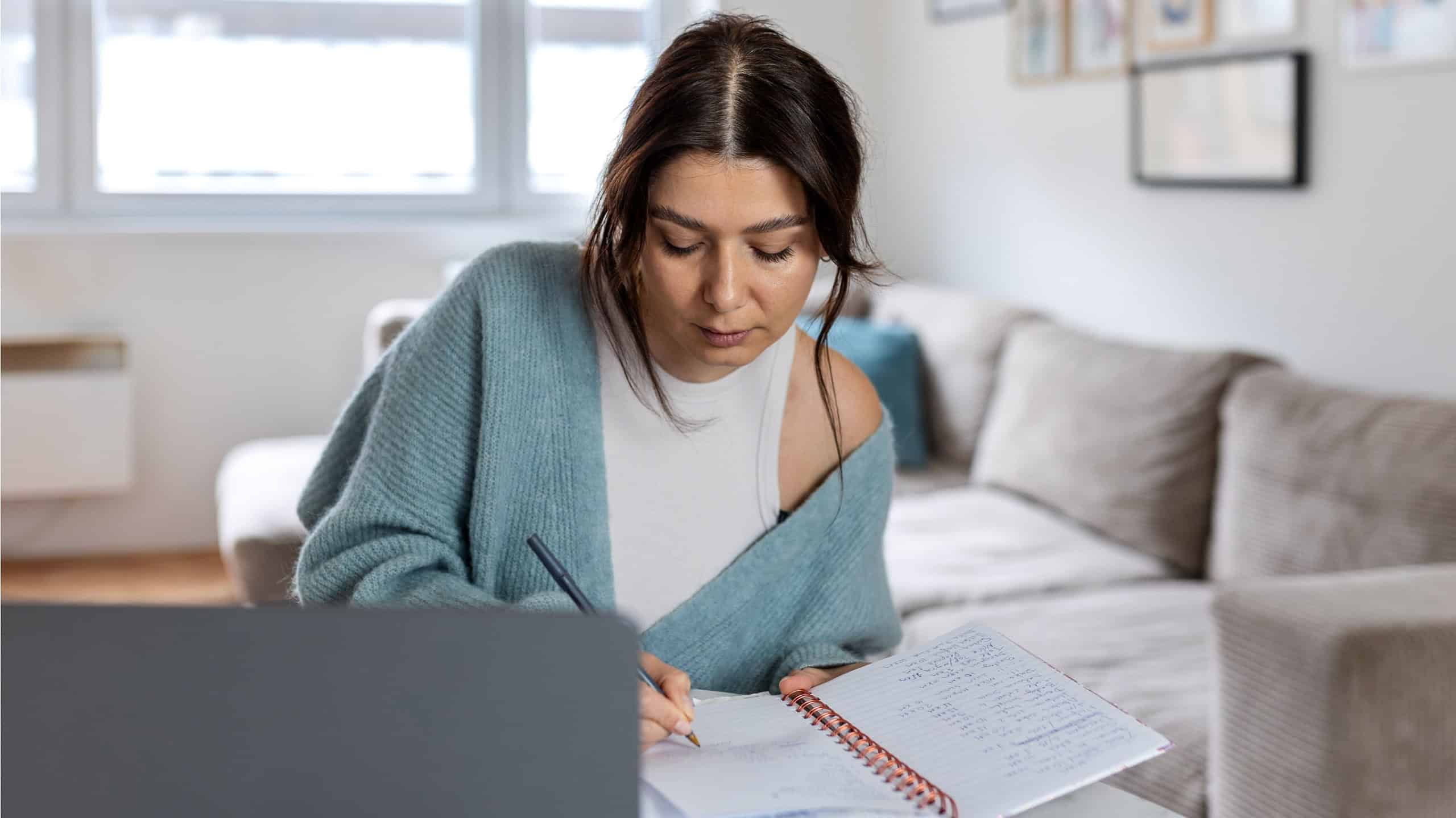 Remote Project Management and Strategic Planning. Focused Female Professional Writing in Notebook while Working on Laptop in Bright Modern Home Office.
