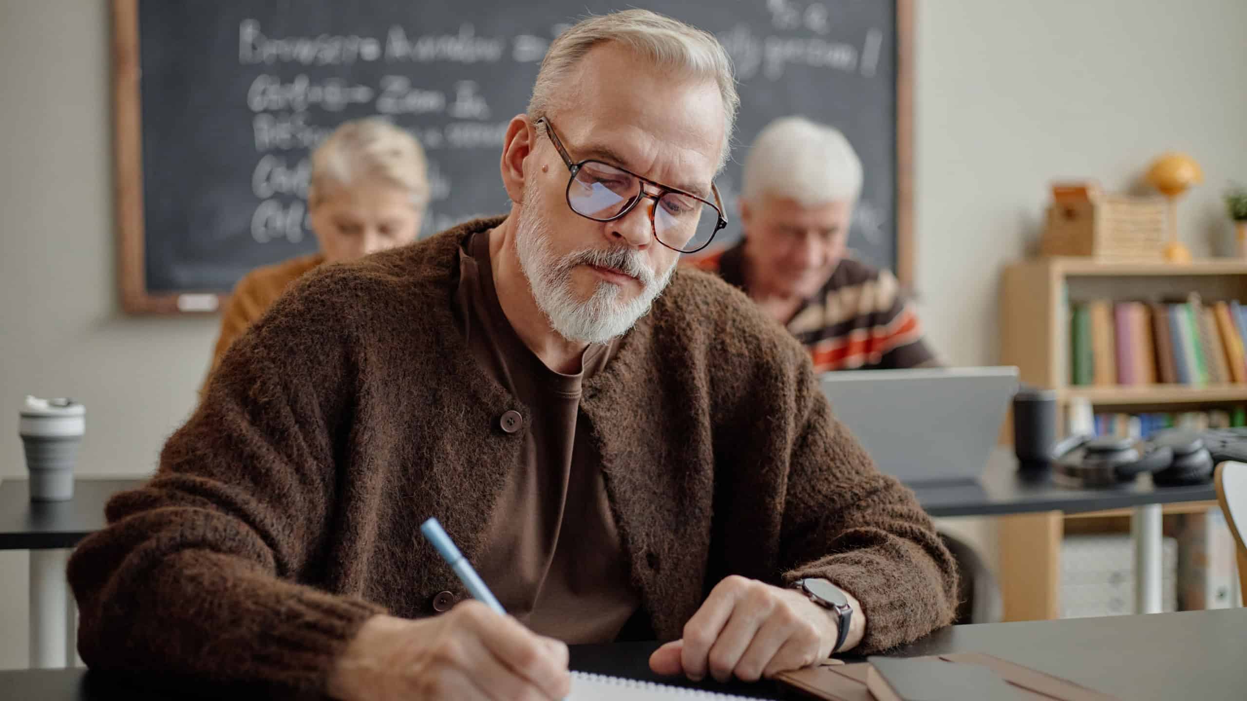 Middle aged Caucasian man wearing glasses sitting at desk, writing in notebook during classroom lesson with senior Caucasian woman and senior Caucasian man studying in background