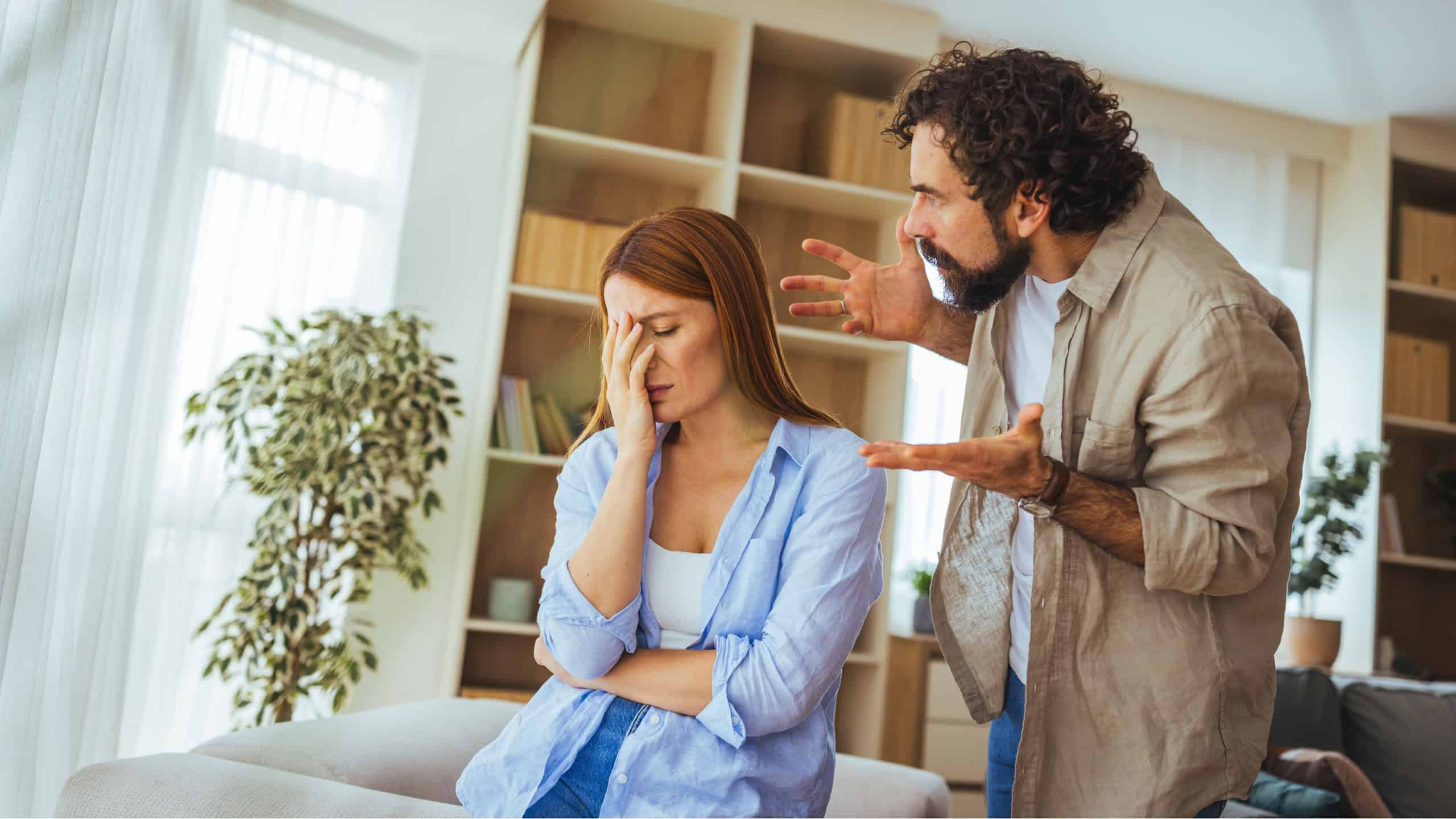 A tense couple argues in a living room as the woman covers her face in frustration and the man gestures angrily, conveying relationship conflict, emotional stress and domestic tension.
