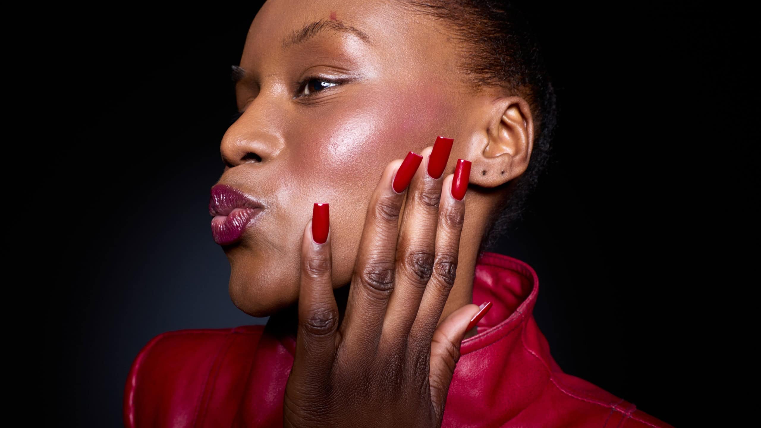 Close-up of a confident black woman with dark skin wearing a red leather jacket and red nail polish. She touches her face with manicured nails against a black background, beauty concept.