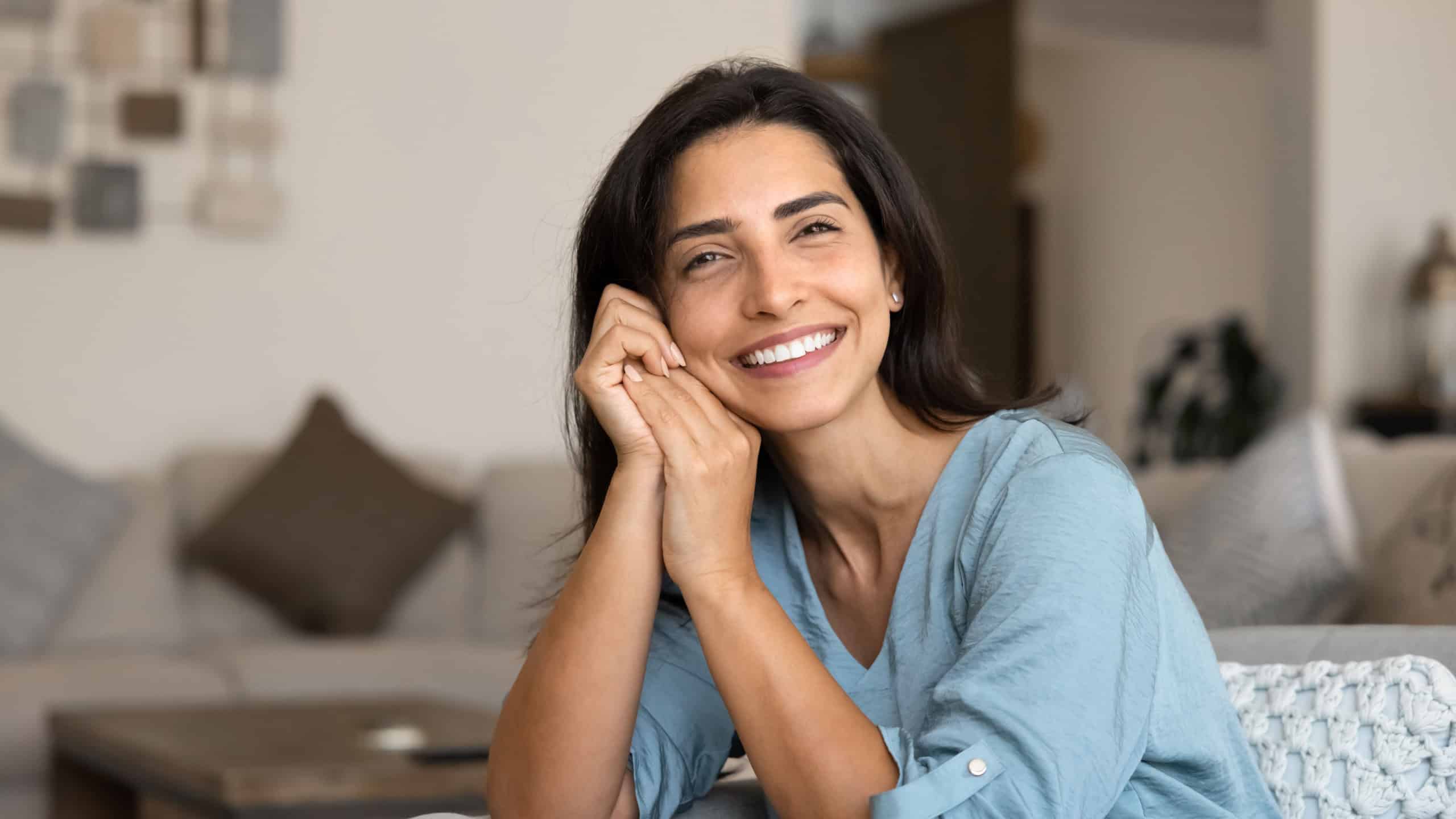 Calm Latina woman sitting comfortable on cozy sofa, looks at camera with contented smile, enjoying weekend rest in fashionable living room, spend weekend, daydreaming indoors. Homeowner lady portrait