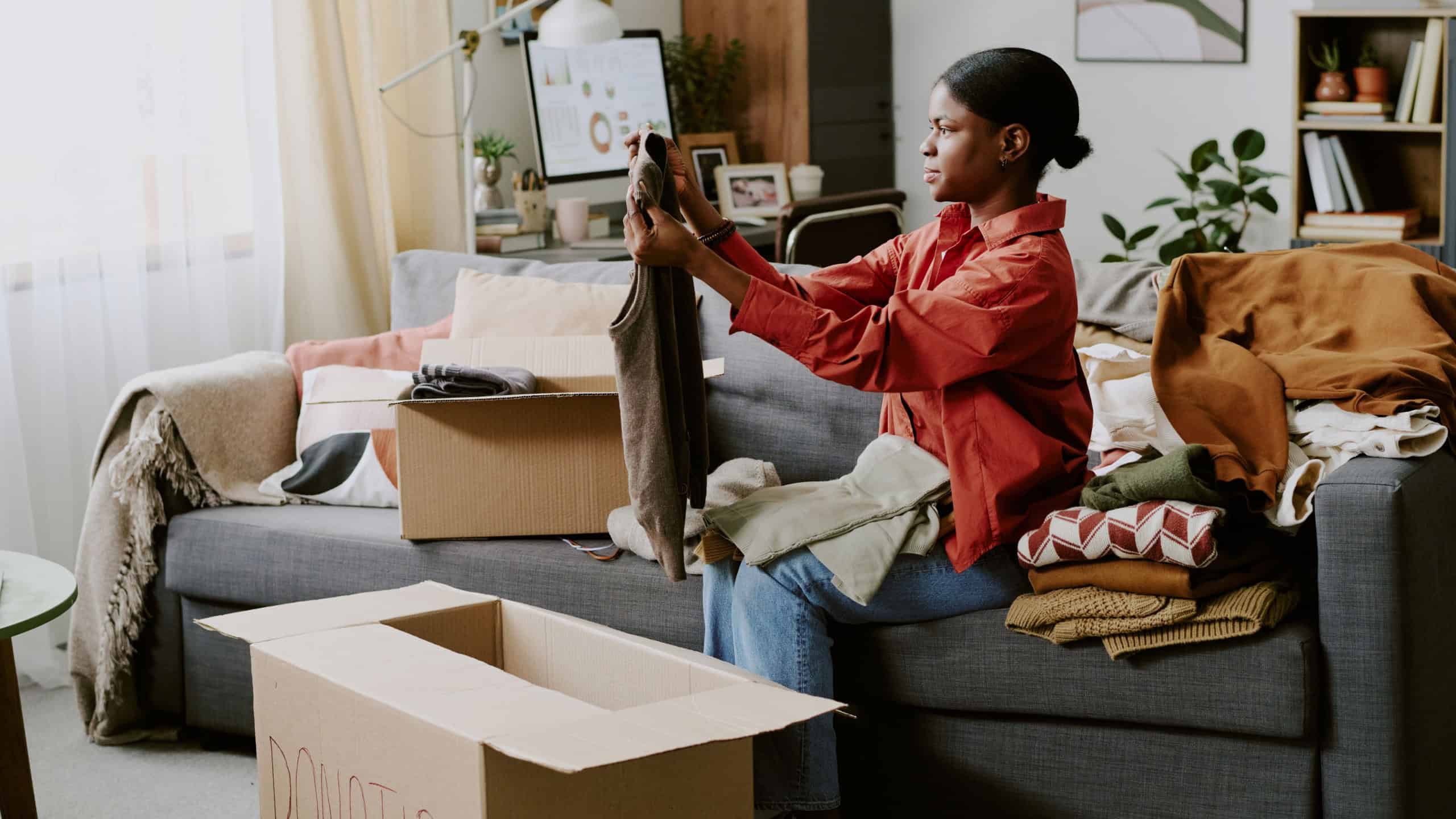 African American young woman sorting folded clothes into donation boxes at home, supporting decluttering and charitable giving. Ideal for nonprofit campaigns, resale apps, sustainability marketing