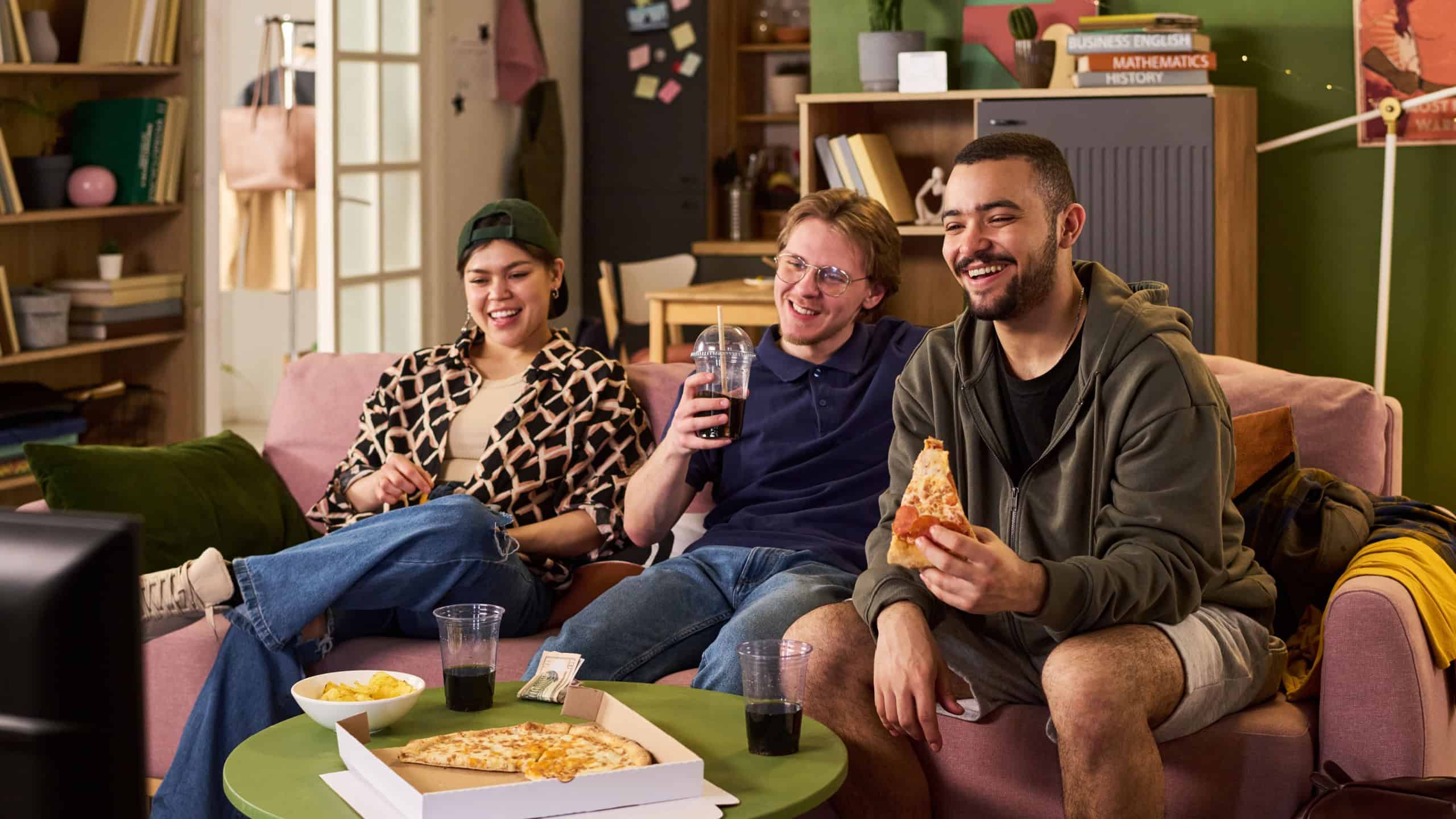 Multiethnic college students relaxing on couch, watching screen, eating pizza, drinking soda, expressing friendship and shared downtime. Ideal for student lifestyle, youth marketing, campus living