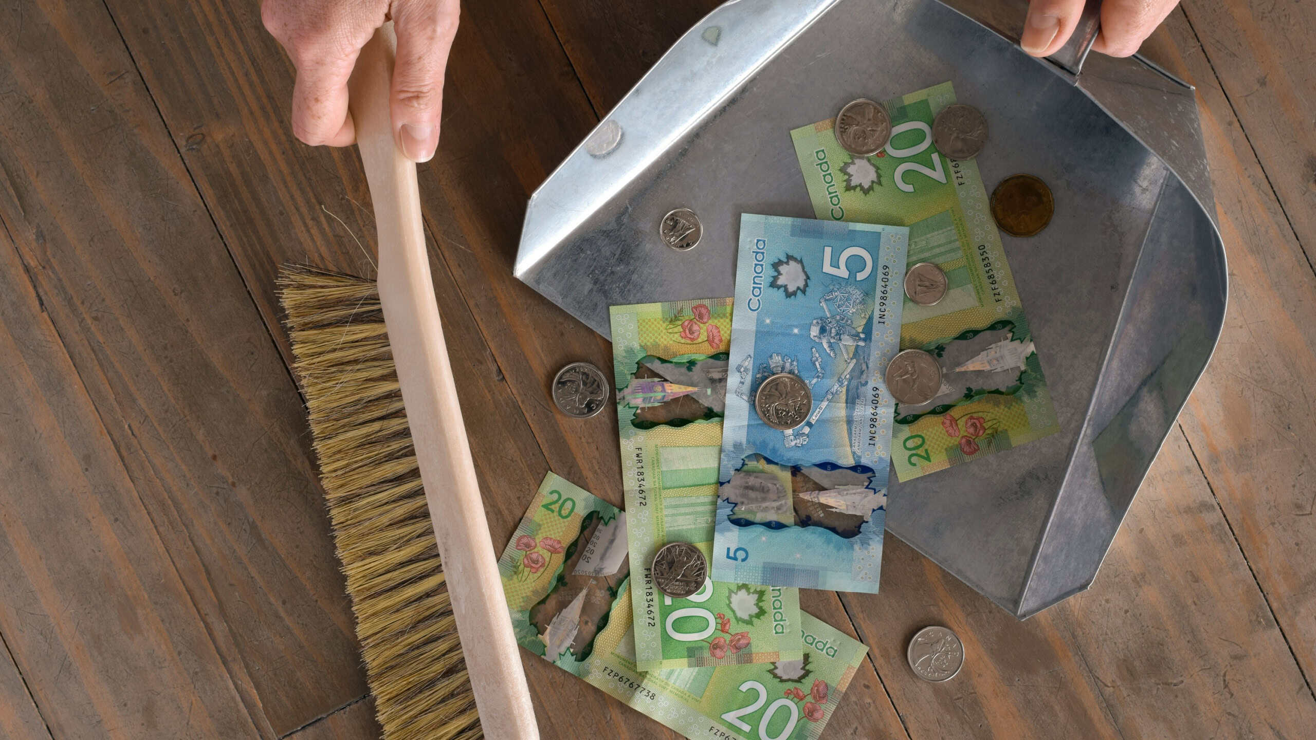 Hands sweeping Canadian dollar bills from floor into dustpan, symbolizing financial loss, rising expenses, debt, or cost of living pressure. Concept image for inflation, recession, taxes, or money management