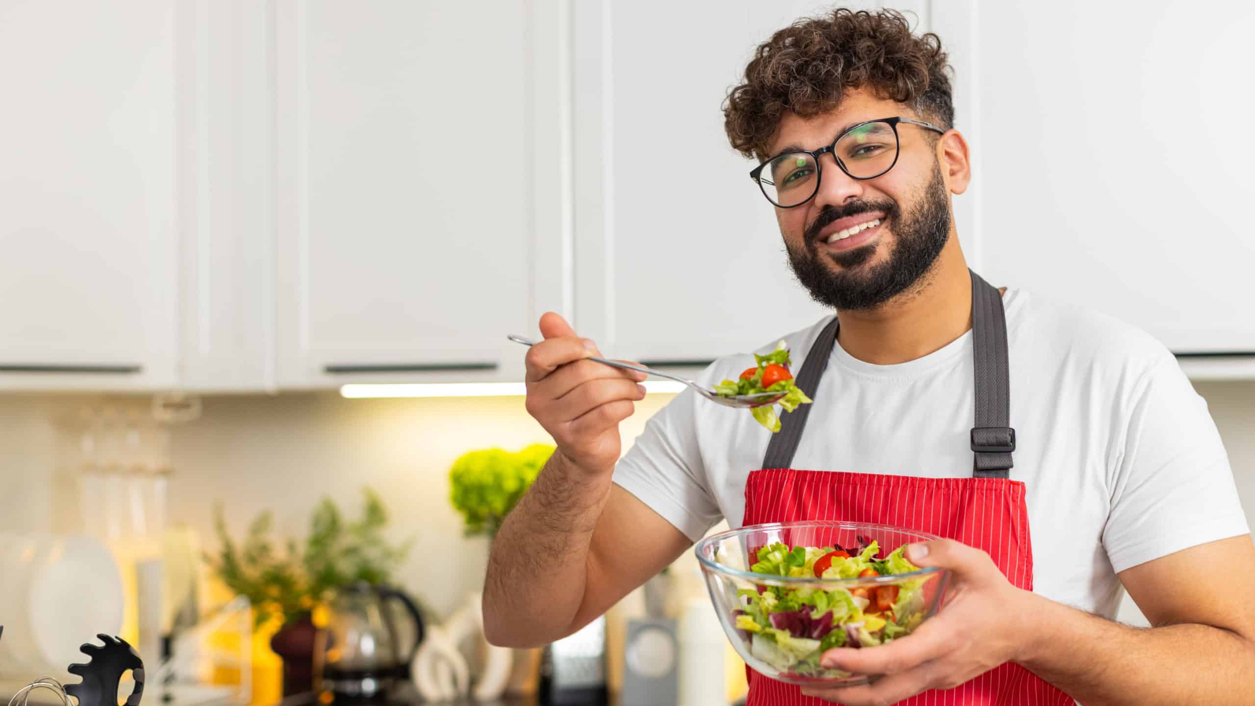 Arabian man tastes fresh salad in kitchen after slicing tomatoes for keto diet and clean nutrition. Young guy feels satisfied and proud choosing healthy eating for fitness at home apartment indoors.