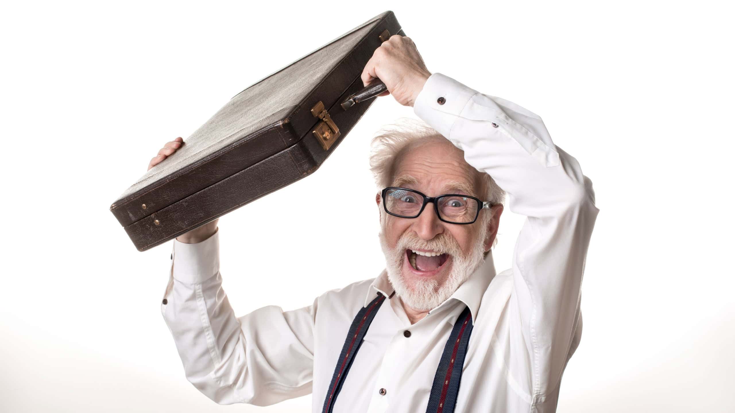 Waist up portrait of delighted old male holding suitcase above his head, expressing excitement. Isolated on background