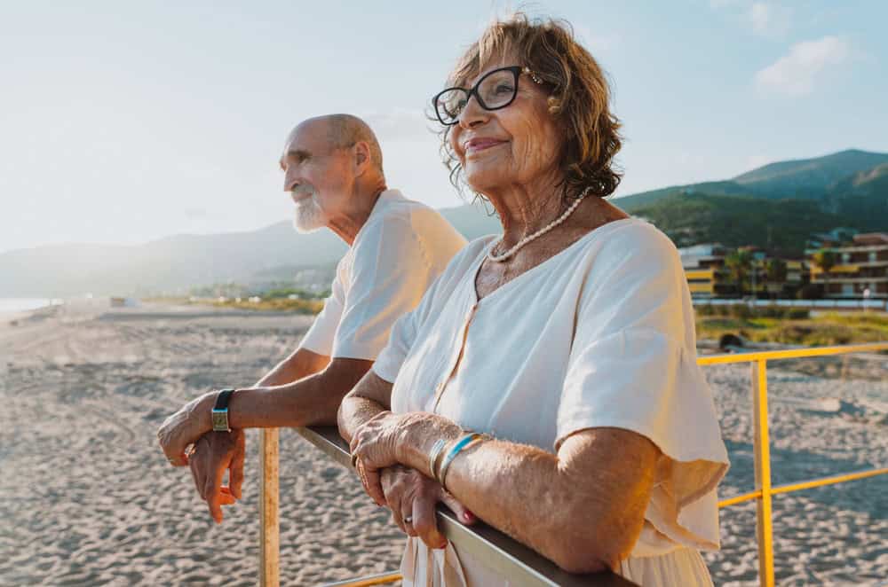 Elder man and woman standing together on a bright yellow lifeguard tower, looking out at the sunny beach and enjoying their active retirement lifestyle